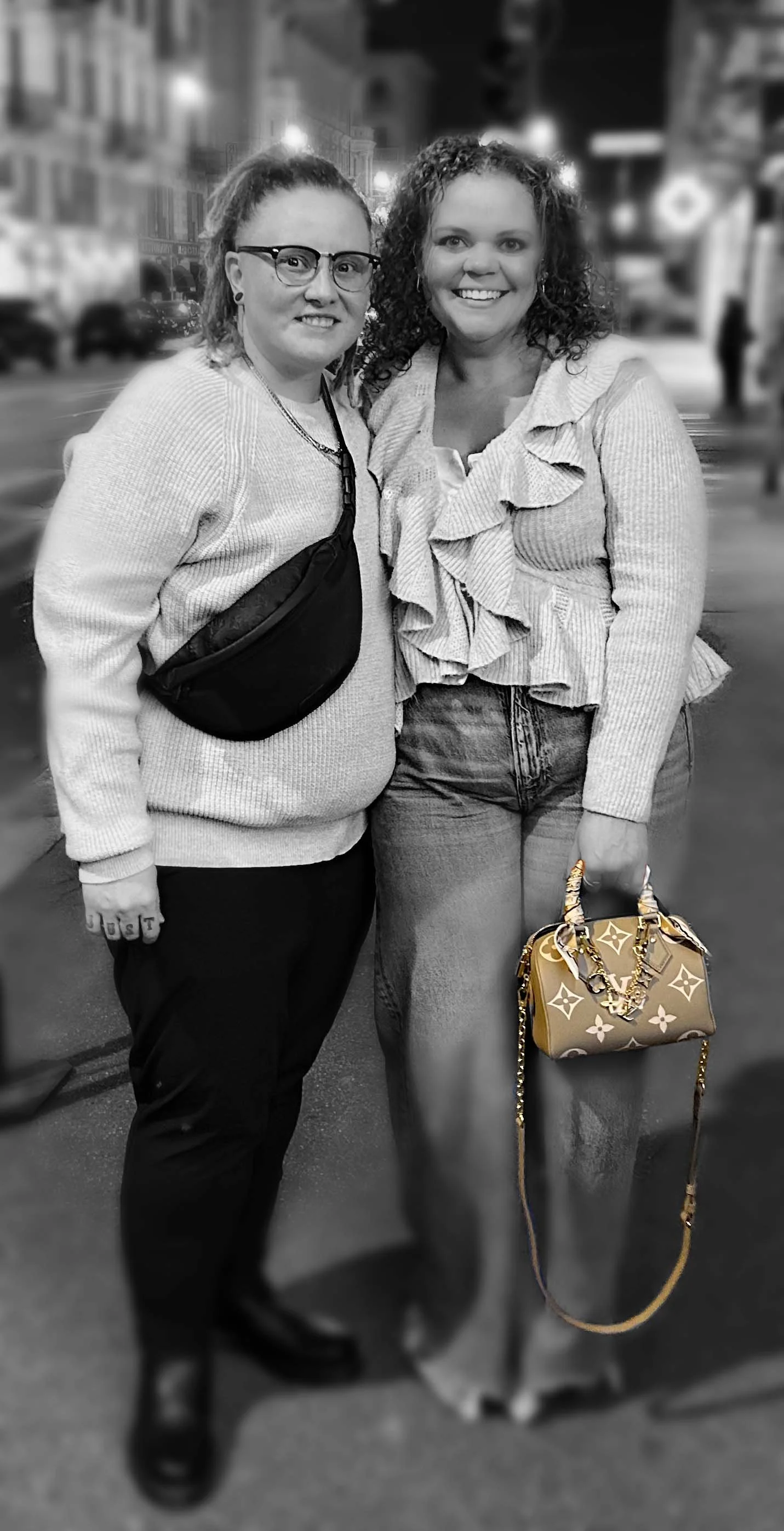 Two women standing together on a city street at night, smiling, with blurred city lights and buildings in the background.