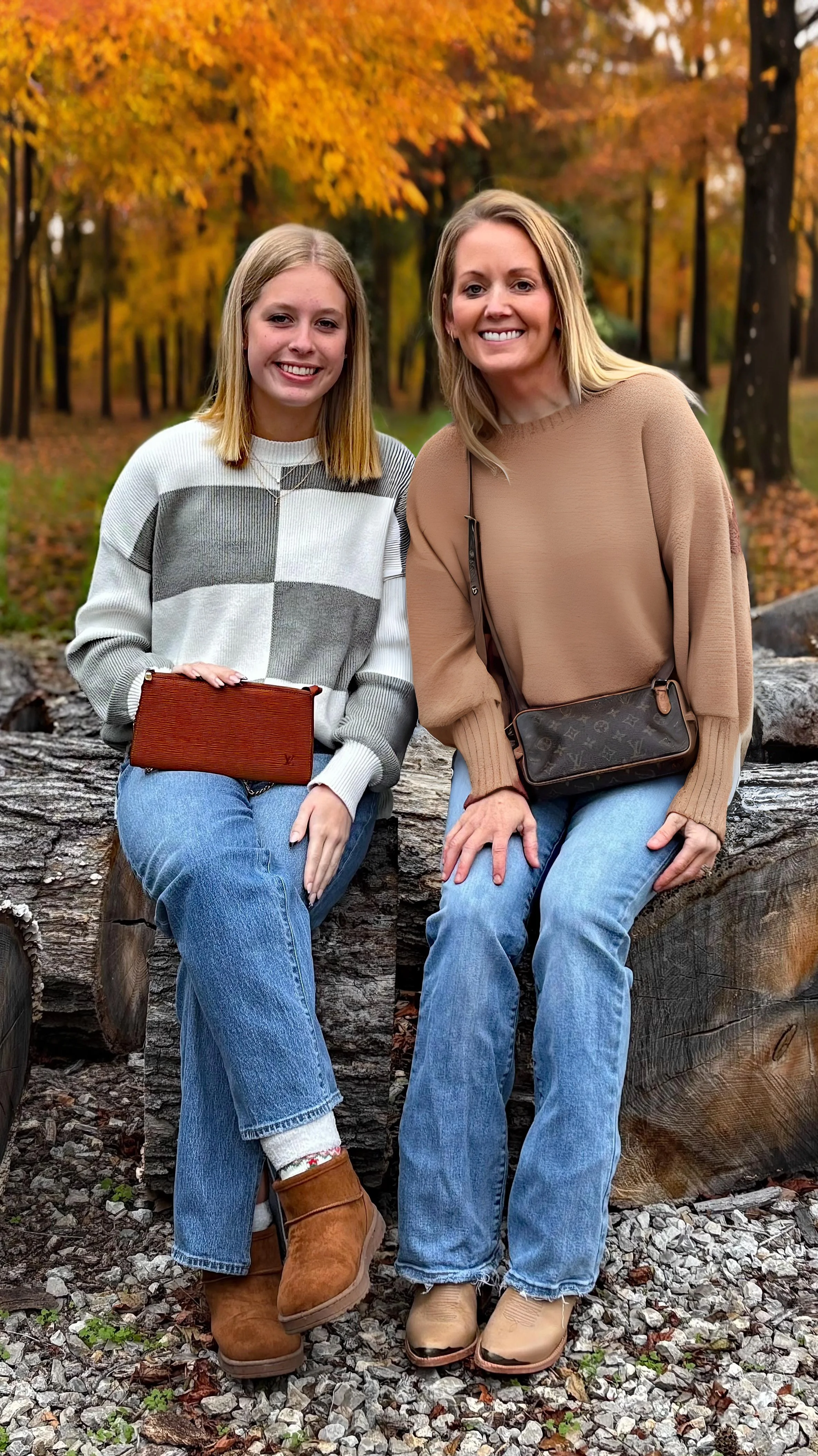 A woman and a girl sitting on a large log in a park with fall foliage in the background. They are smiling and wearing casual autumn clothing.