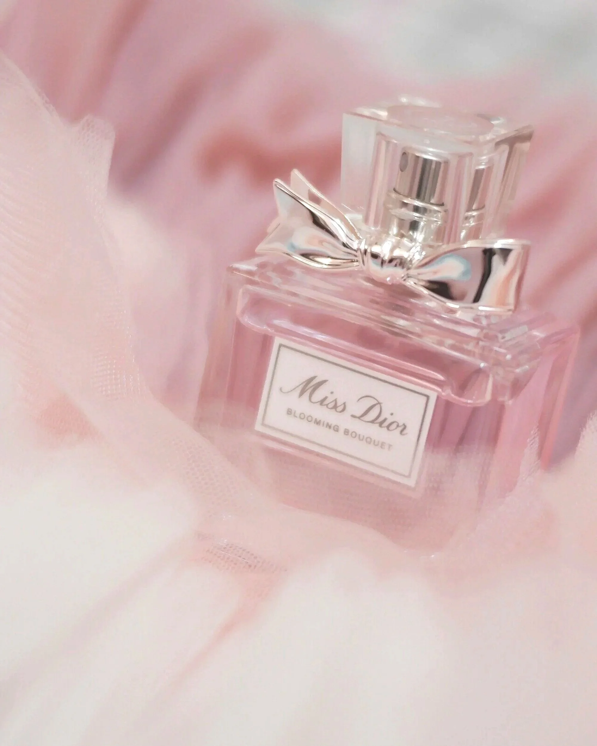 A bottle of Miss Dior Blooming Bouquet perfume with a silver bow, resting on pink fabric.