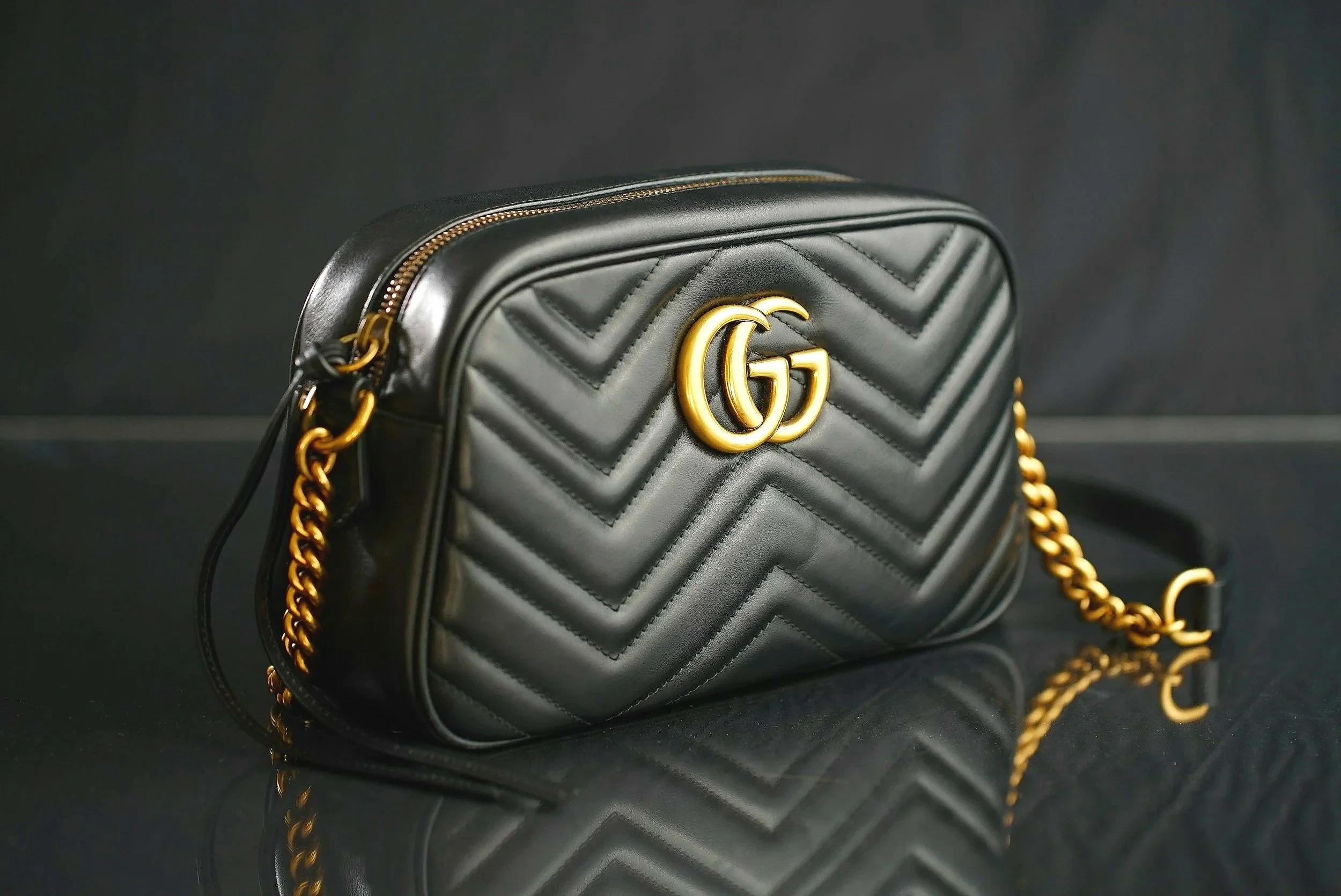 Black quilted leather handbag with gold GG logo and chain strap, on a reflective black surface.