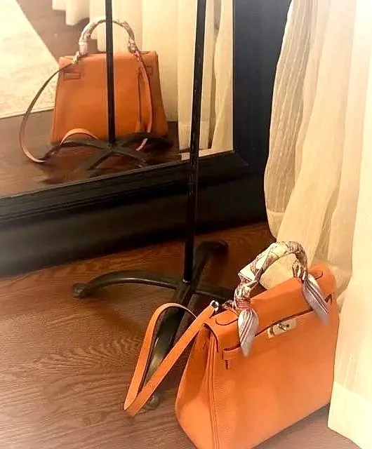 An orange handbag with a patterned scarf tied to its handle, placed on the floor near a mirror and a curtain.