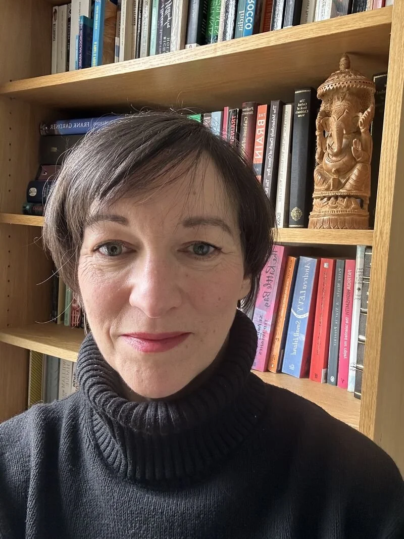 Shifra Cook. A woman with short dark hair and blue eyes, wearing a black turtleneck, stands in front of a wooden bookshelf filled with books and a carved wooden statue of Ganesha.