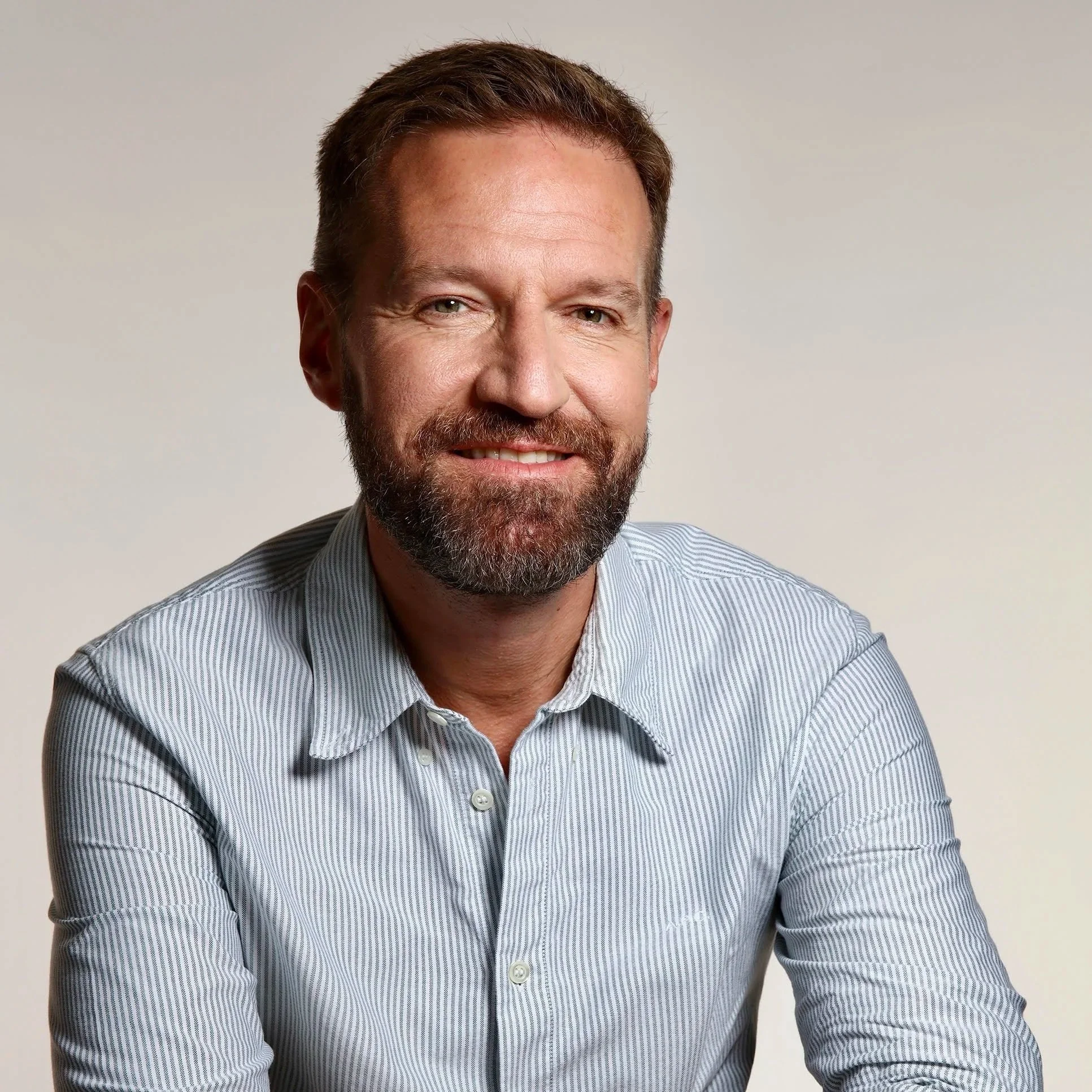 Richard Chapman of Richard Chapman Studies. A smiling man with a beard in a light blue striped button-up shirt against a neutral background.