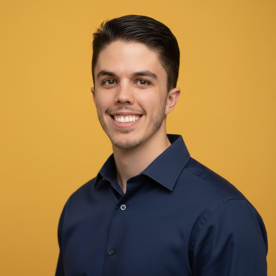 A young man with dark hair, wearing a navy blue shirt, smiling at the camera against a yellow background.