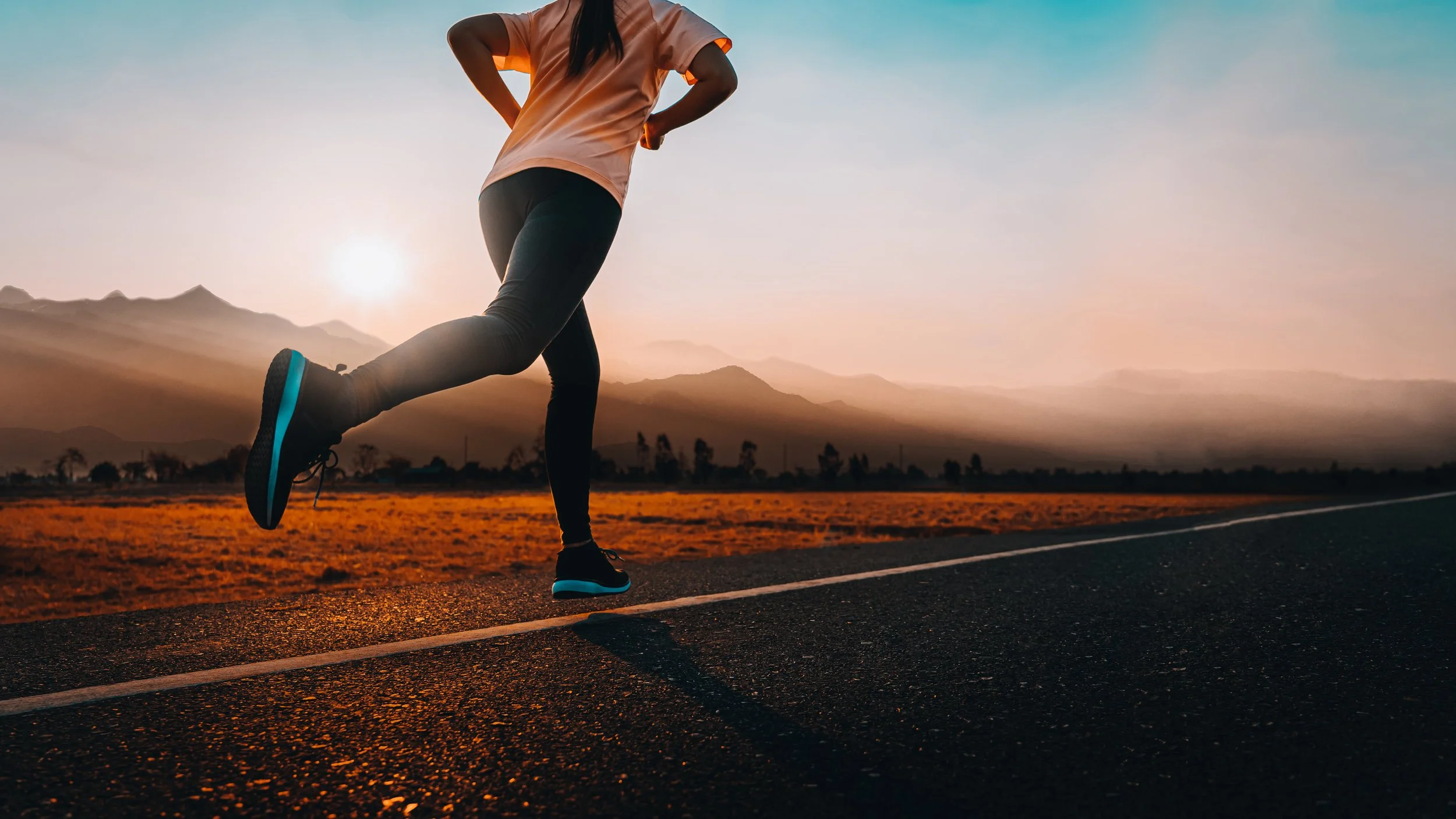 A person running on a road during sunrise or sunset with mountains in the background, wearing a light pink shirt and black leggings.