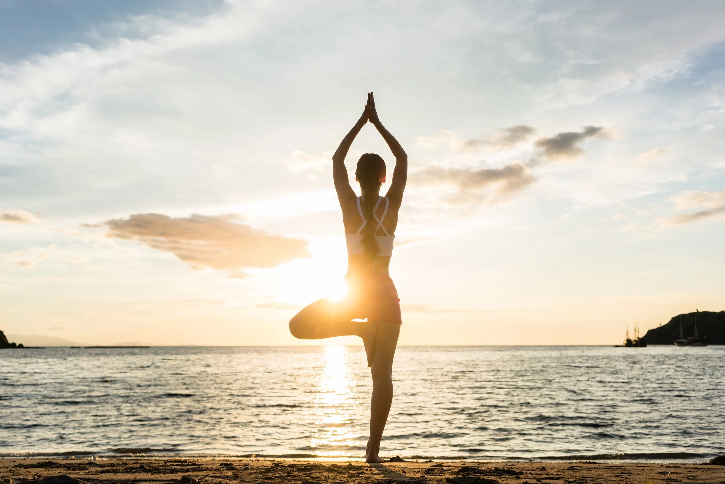 A woman practicing yoga in a tree pose on the beach at sunset, with ocean and cloudy sky in the background.