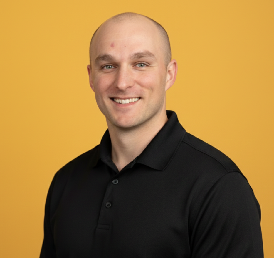 A smiling man with a shaved head wearing a black collared shirt against a yellow background.