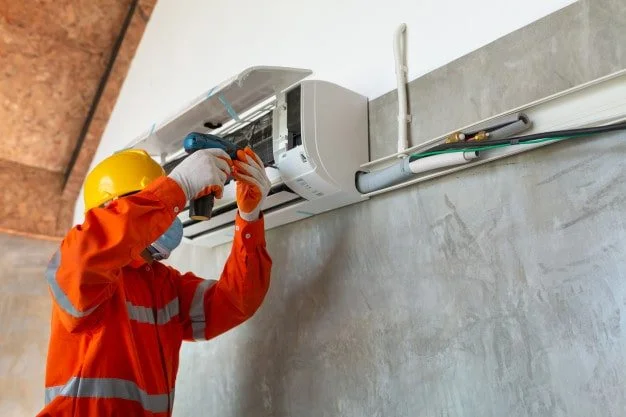 Bir elektrikçi, duvardaki klimaların elektrik bağlantılarını tamir ederken şapka ve maske takıyor.