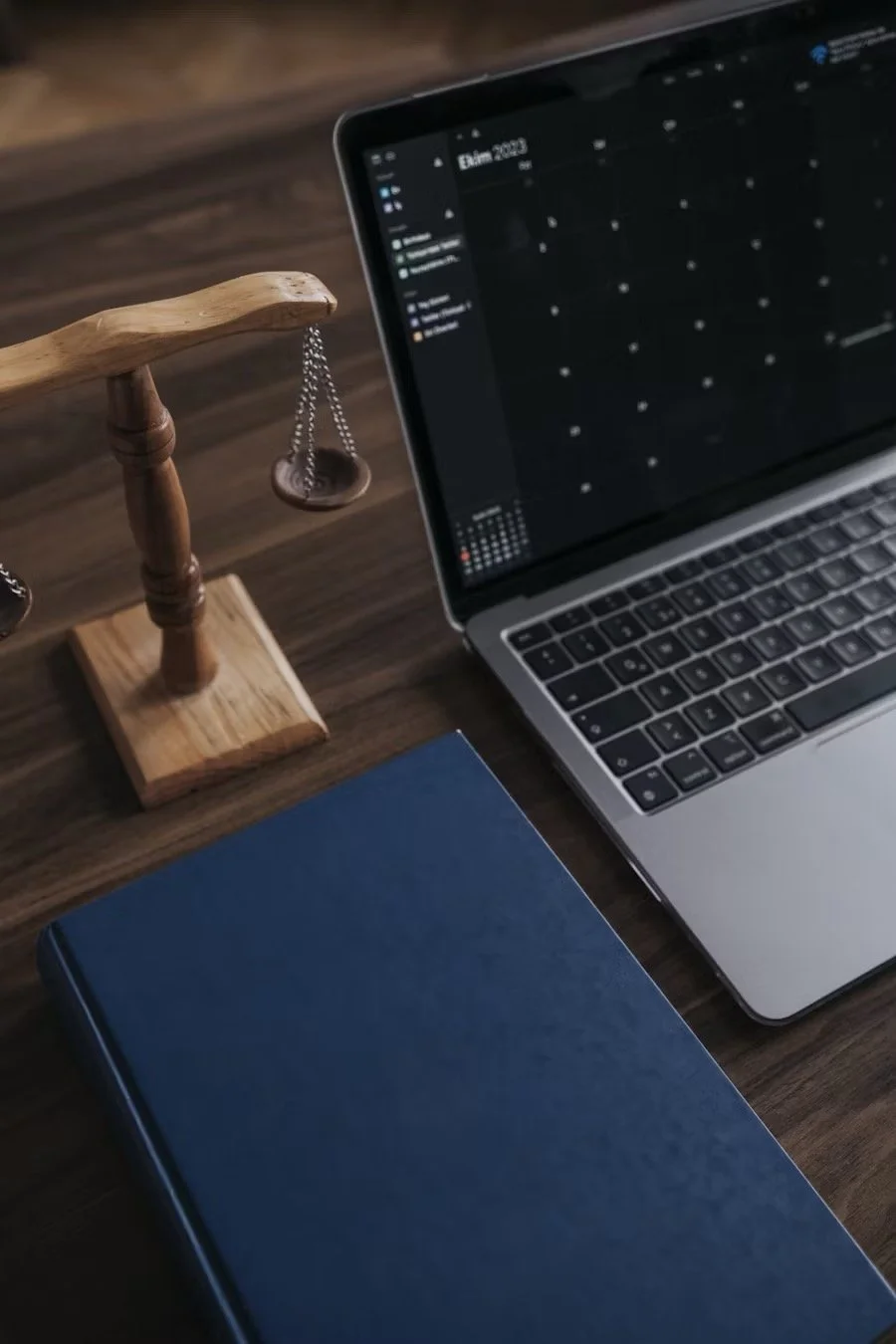 A wooden desktop with a laptop displaying a calendar app, a closed blue notebook, and a small wooden balance scale.