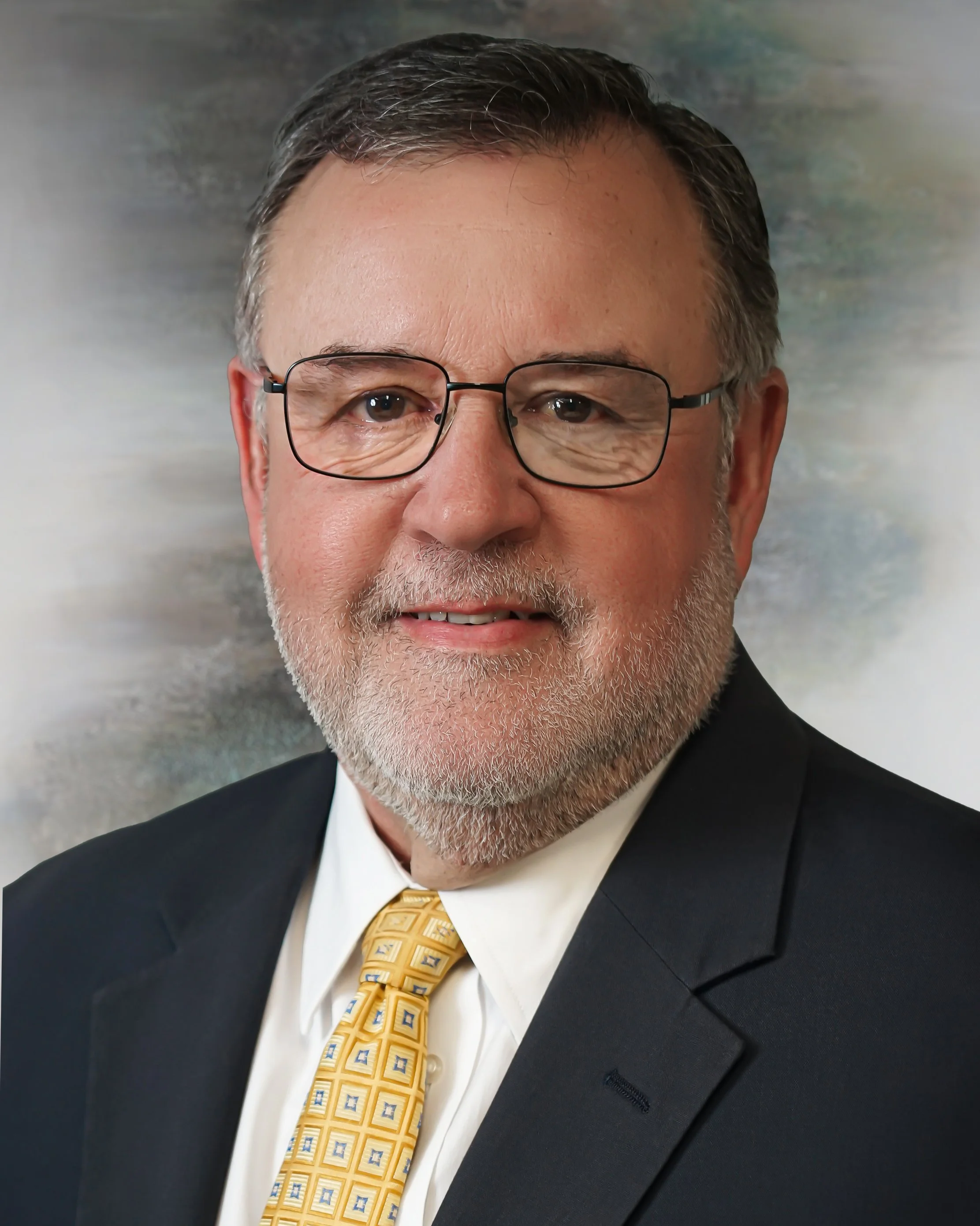 Professional headshot of a middle-aged man with glasses, gray hair, and a trimmed beard, wearing a black suit, white shirt, and yellow patterned tie, against a blurred background.