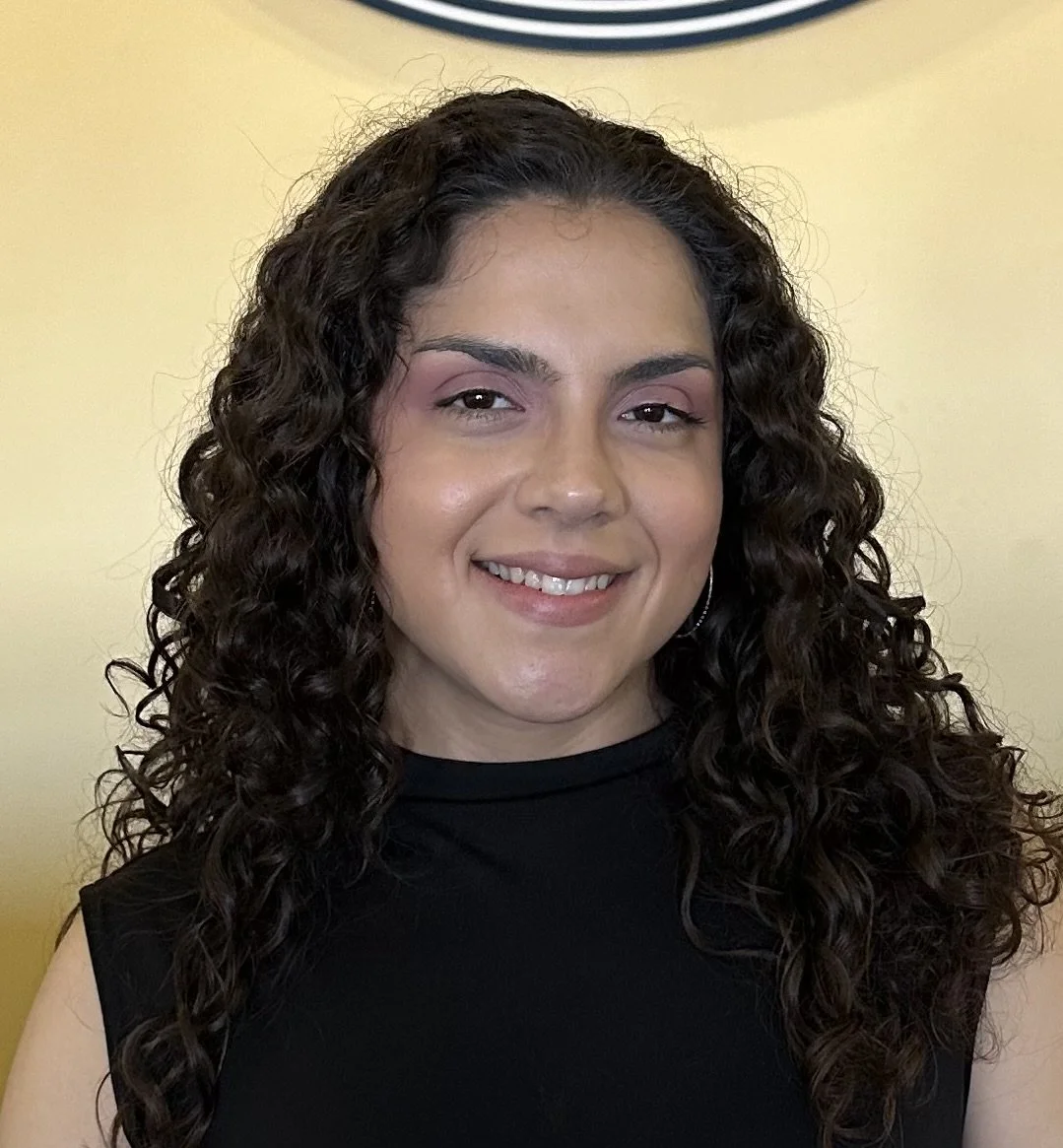 A young woman with long, curly dark hair wearing a sleeveless black top, smiling slightly at the camera, with a light yellow background.