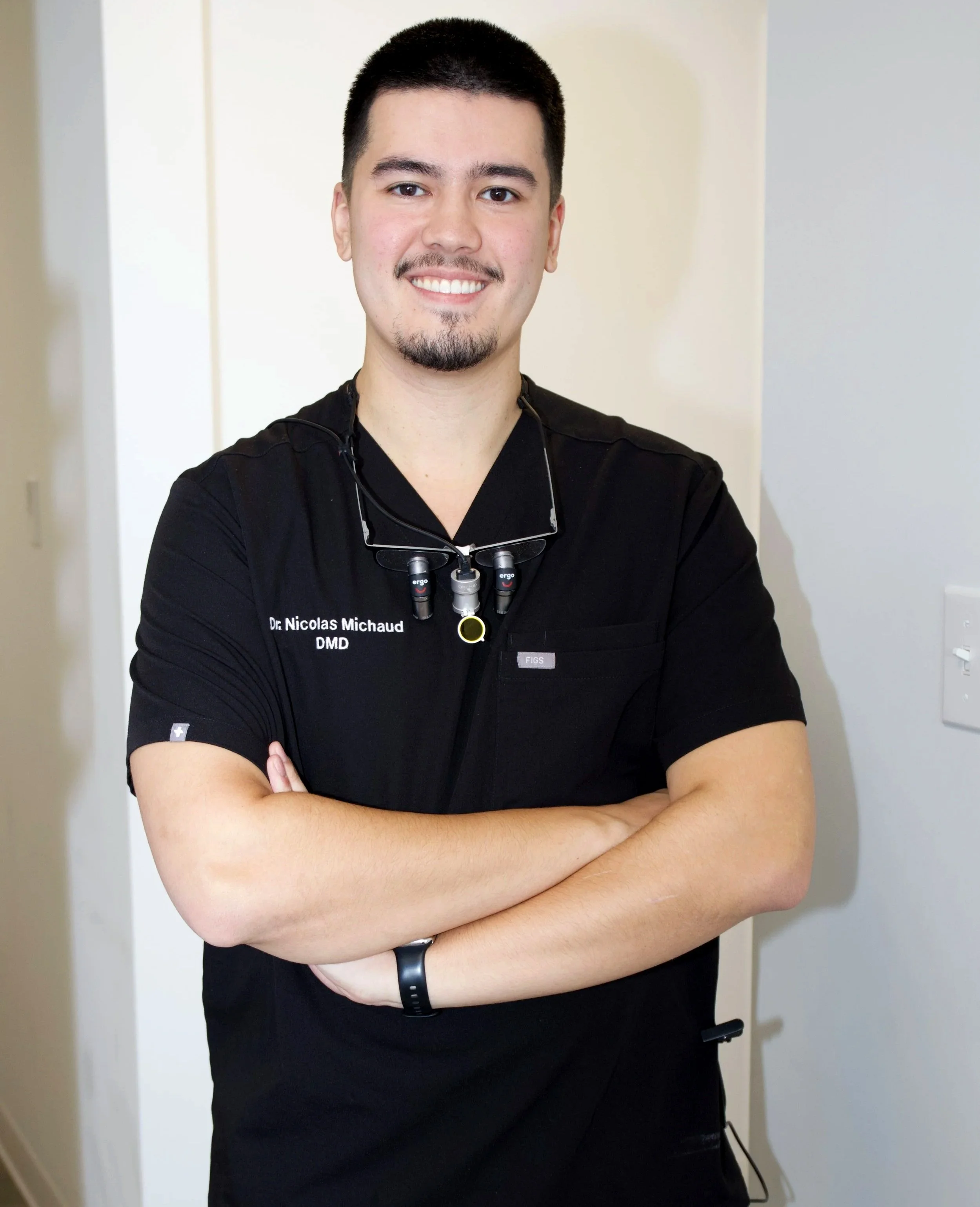 Un homme souriant, portant une tenue médicale noire avec un badge indiquant son nom et diplôme, en cabinet dentaire.