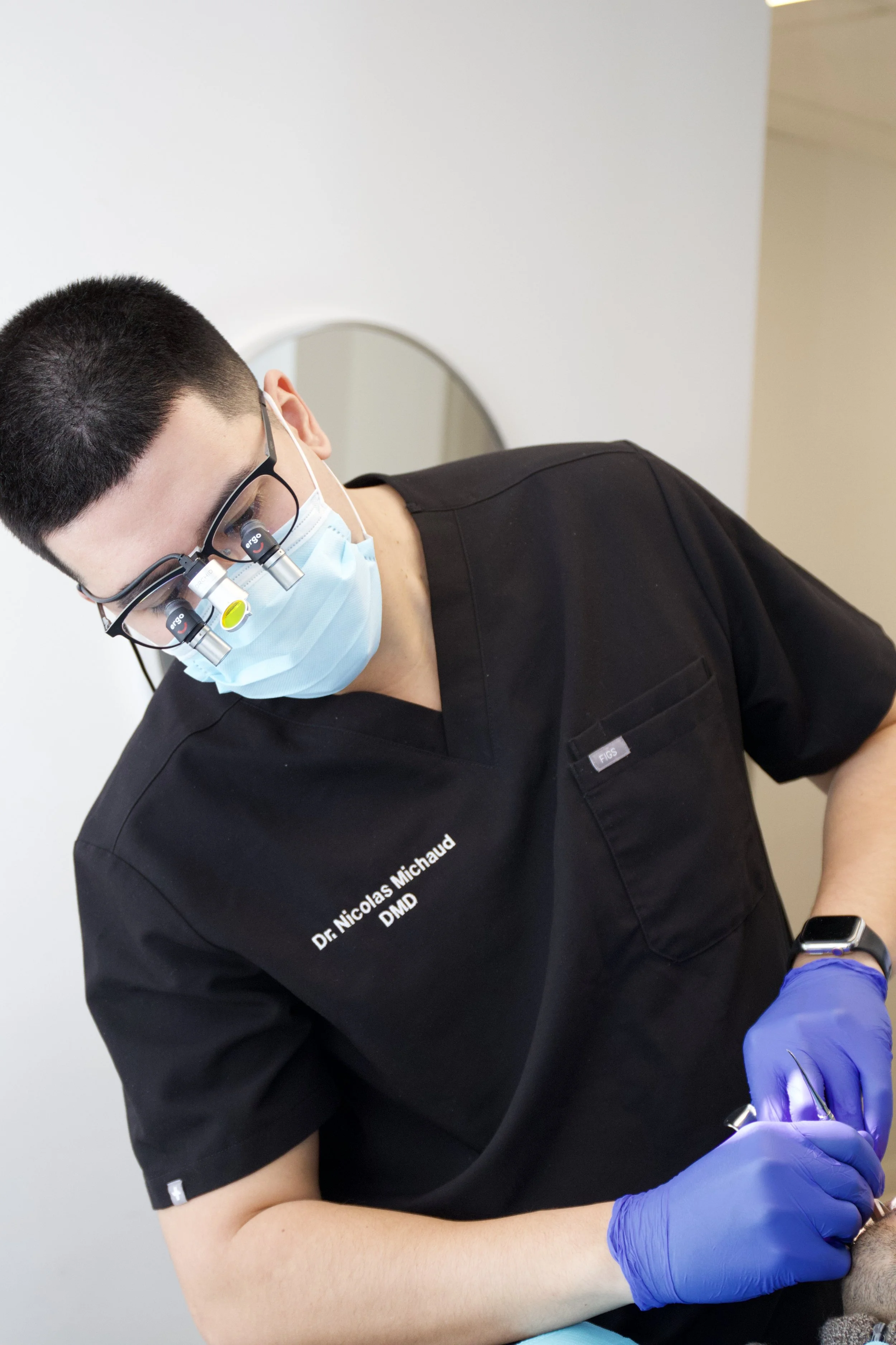 Un dentiste portant une blouse noire, des lunettes avec une loupe, un masque chirurgical, des gants bleus et une montre, effectue un traitement dentaire.
