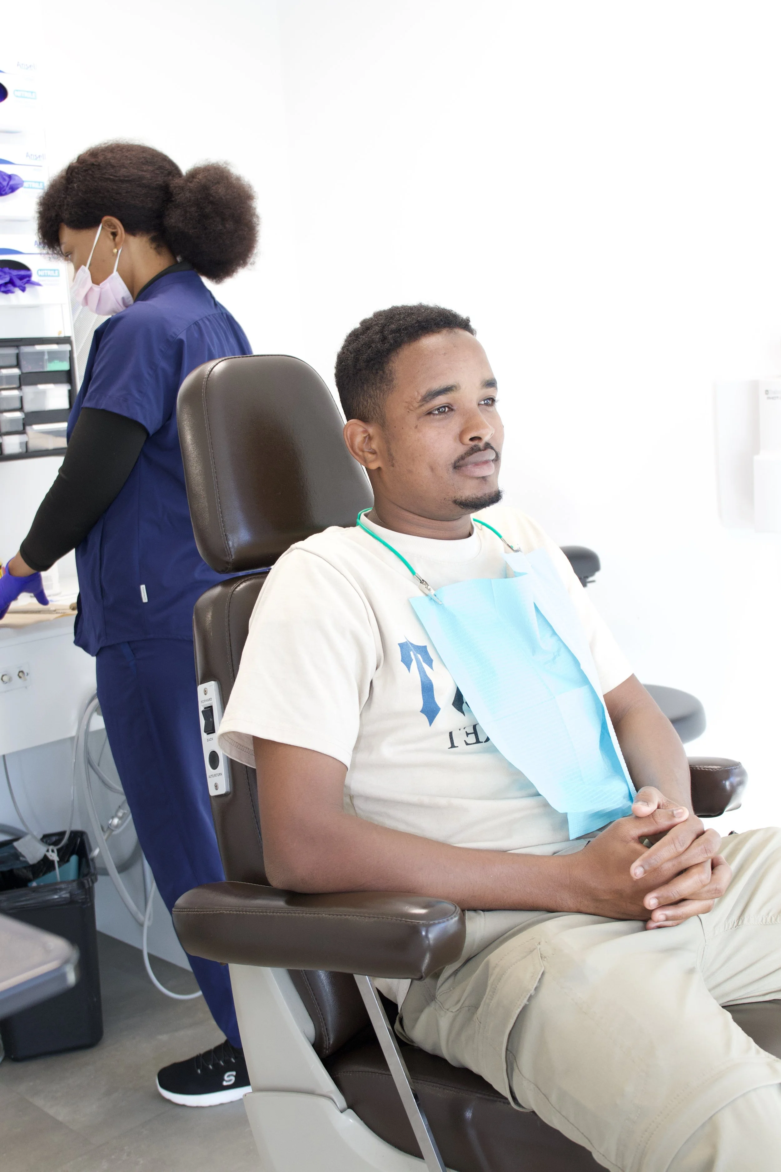 Un homme assis dans un fauteuil médical avec une blouse en papier bleue autour du cou, dans une clinique ou un centre médical, pendant qu'une professionnelle de santé en uniforme bleu et masque examiné des documents ou des échantillons en arrière-plan.