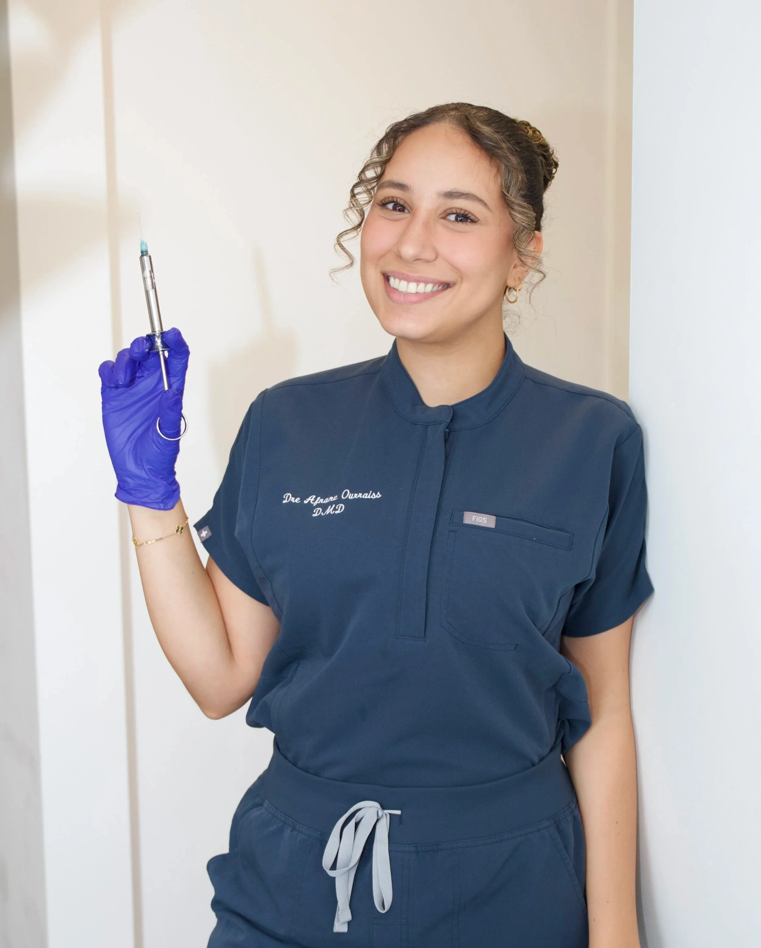 Une infirmière souriante en uniforme bleu, tenant une seringue, dans un environnement médical.