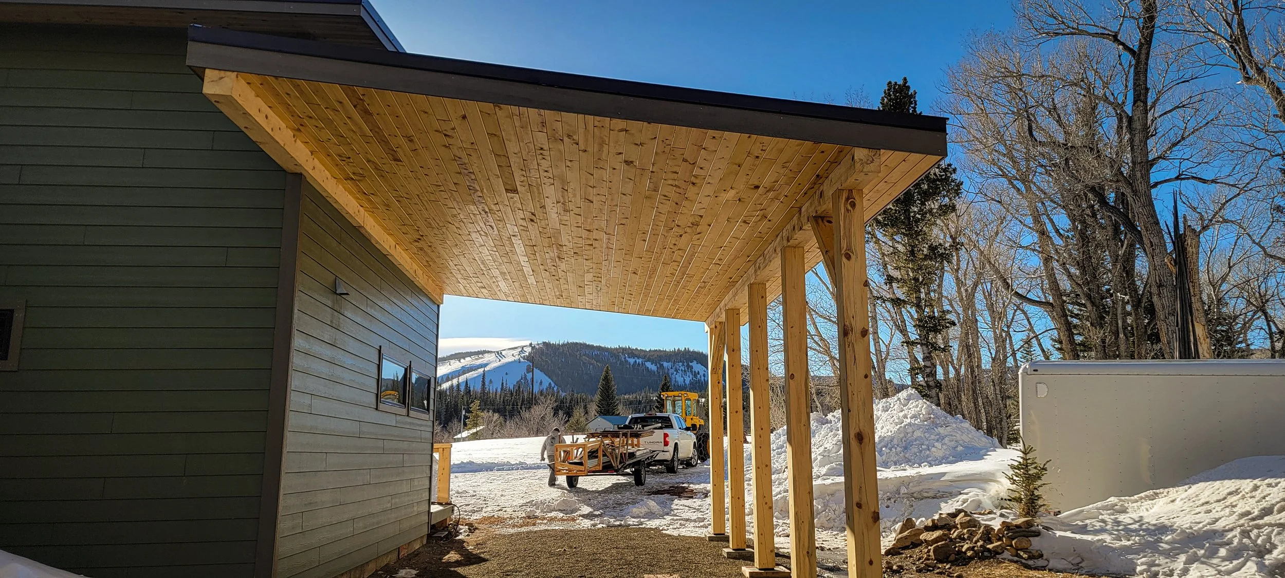 View of a construction site showing a wooden porch extension being built on a house in a snowy landscape, with mountains and trees in the background.