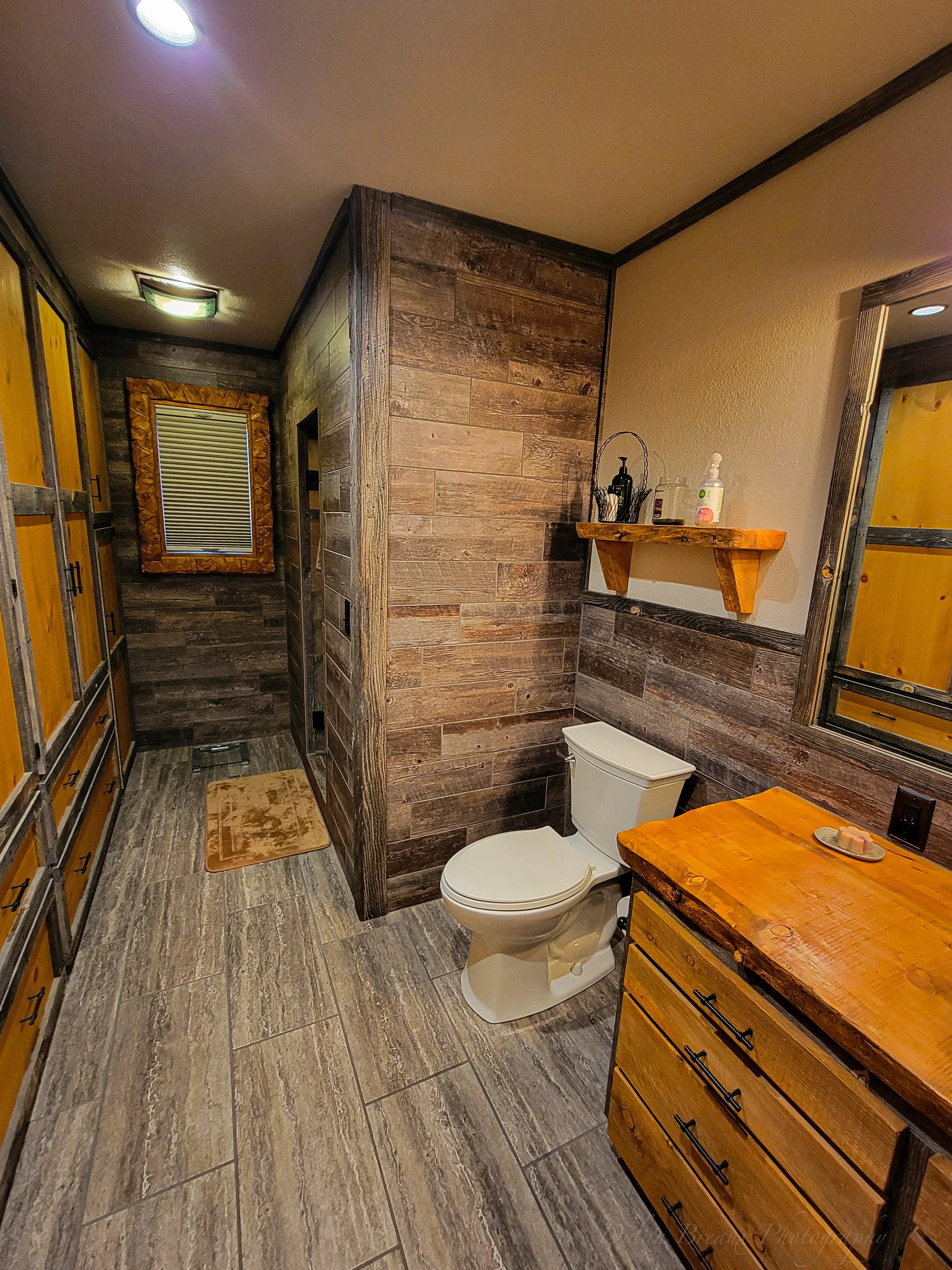 Bathroom with wood-paneled walls, a toilet, a wooden vanity with a matching wooden shelf above it, and a mirror. There are wooden cabinets on the left and tiles on the floor.