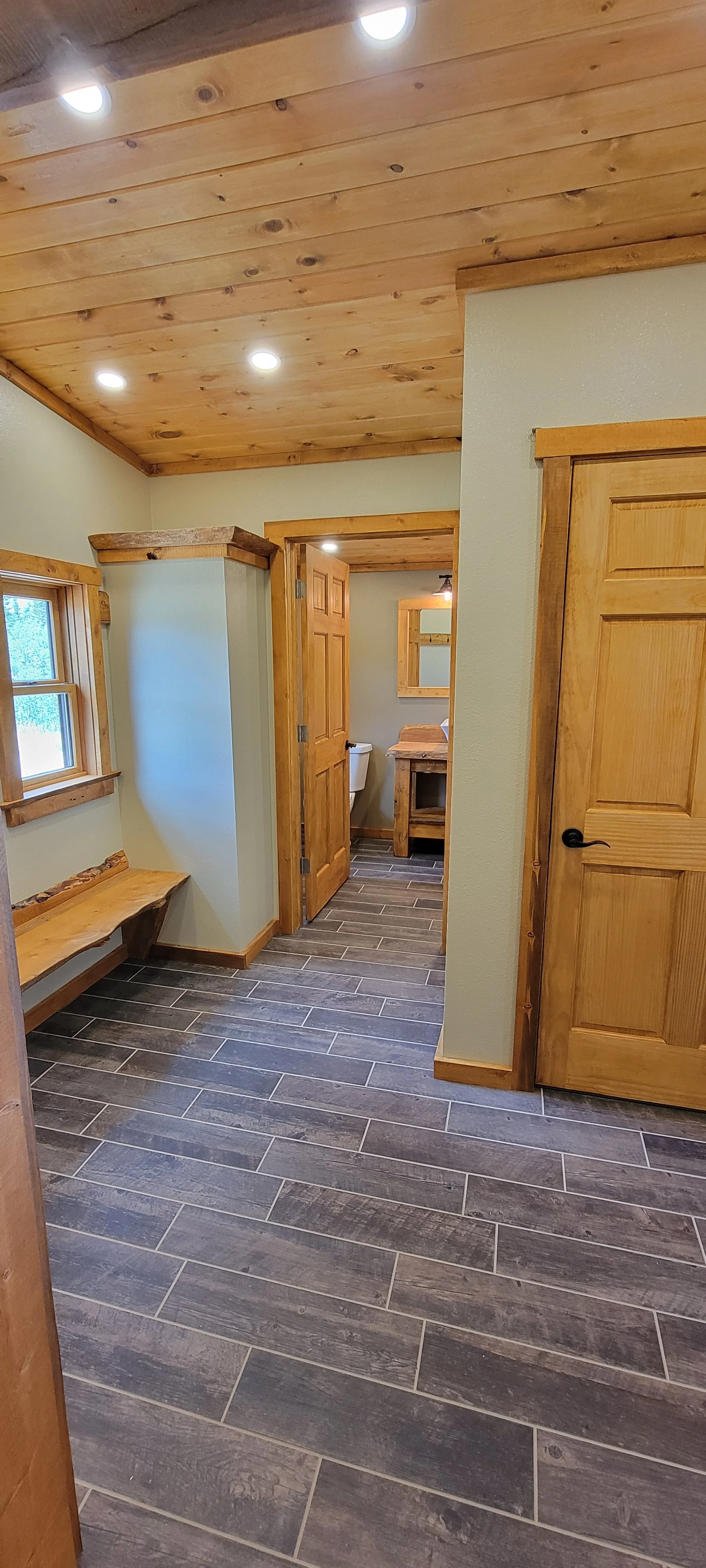 Interior view of a rustic room with wood-paneled ceiling, wooden doors, a bench, and a small bathroom with a sink and mirror.