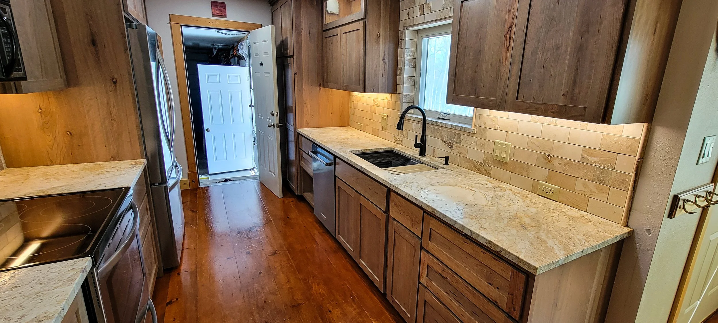 A kitchen with wooden cabinets, a speckled granite countertop, a black sink with a black faucet, a window above the sink, a stainless steel dishwasher, an electric stovetop, and a refrigerator. The floor is wooden, and there is an open door leading outside.
