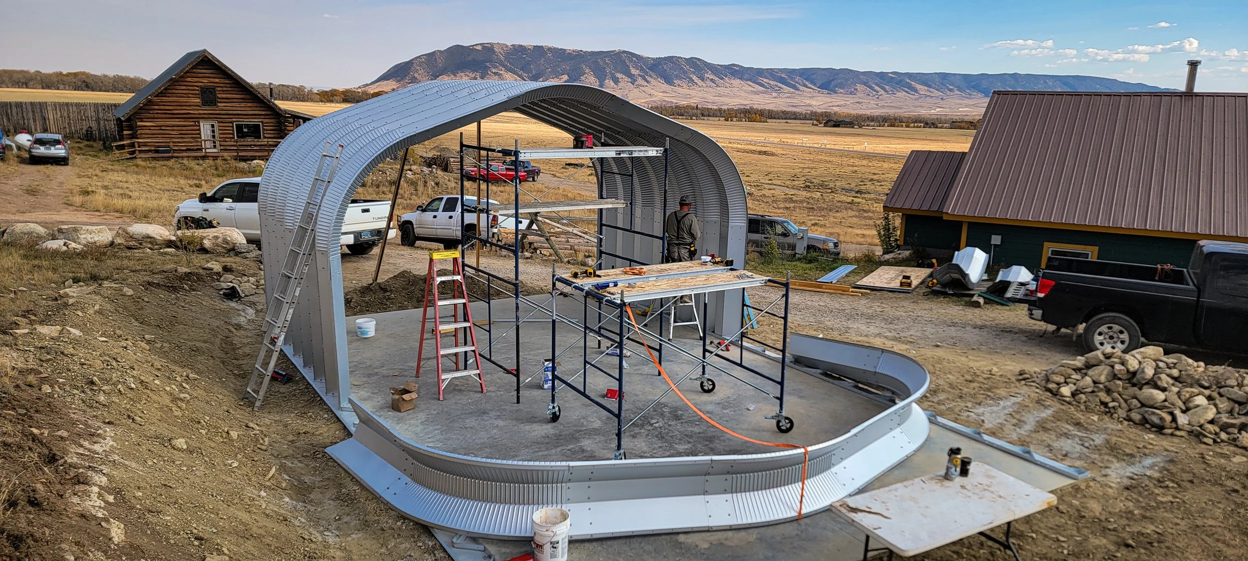 Construction of a curved metal structure on a concrete foundation outdoors with mountains in the background and various tools and scaffolding set up.