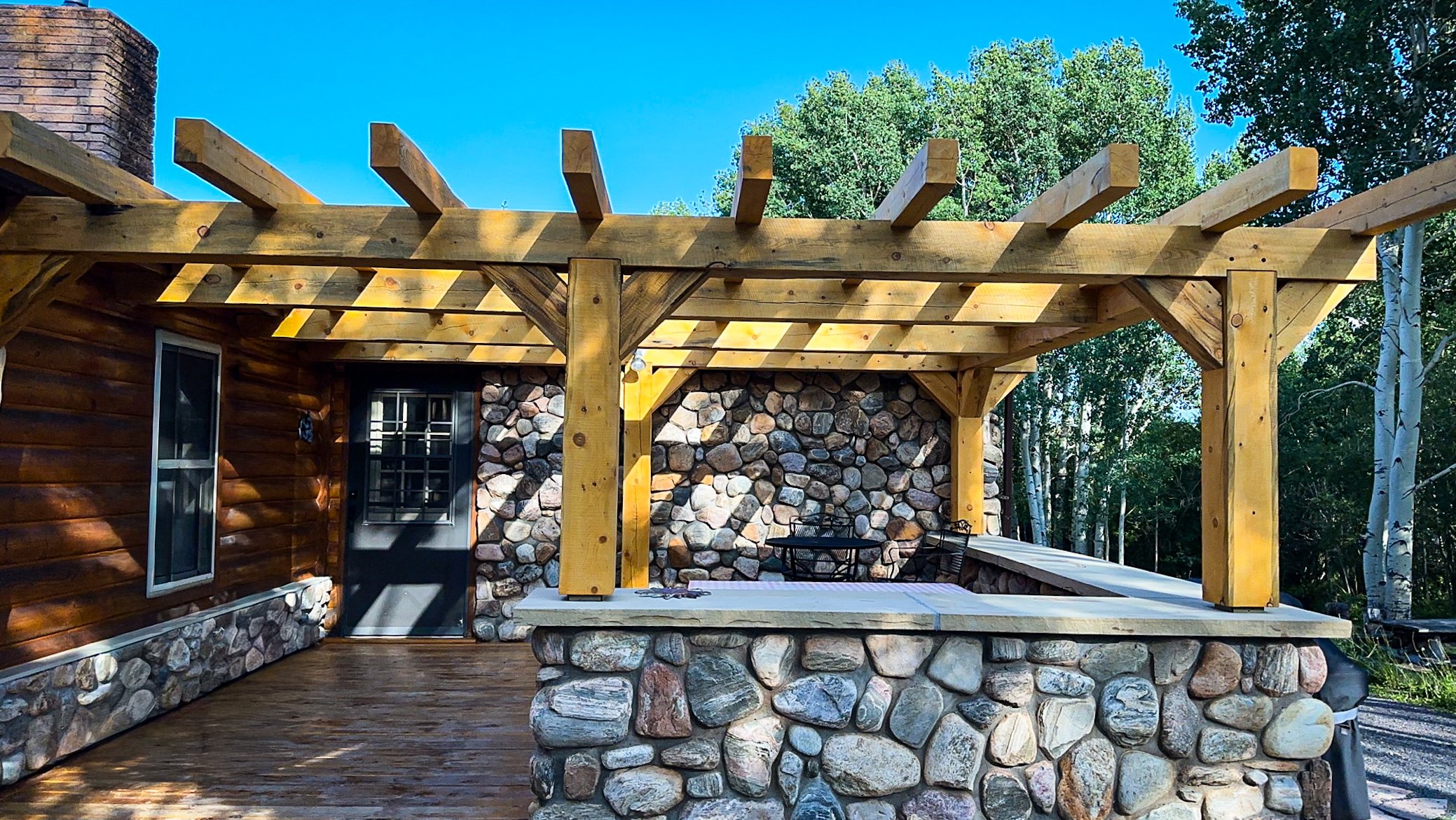 Exterior view of a house with a wooden deck and pergola under construction, surrounded by trees and a clear blue sky.