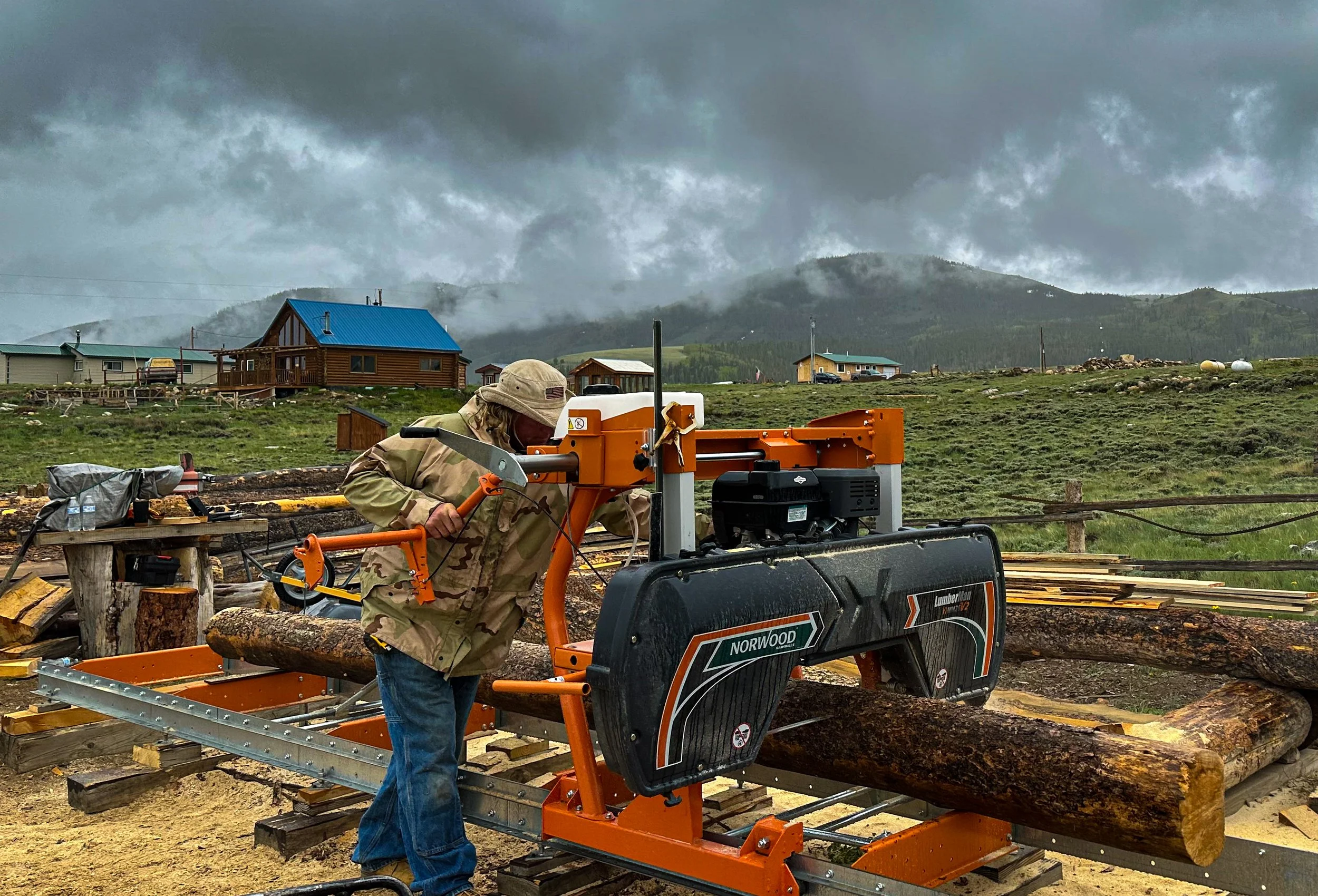A person in camouflage jacket and cap operating a log cutting machine outdoors, with a rural landscape and cloudy mountains in the background.