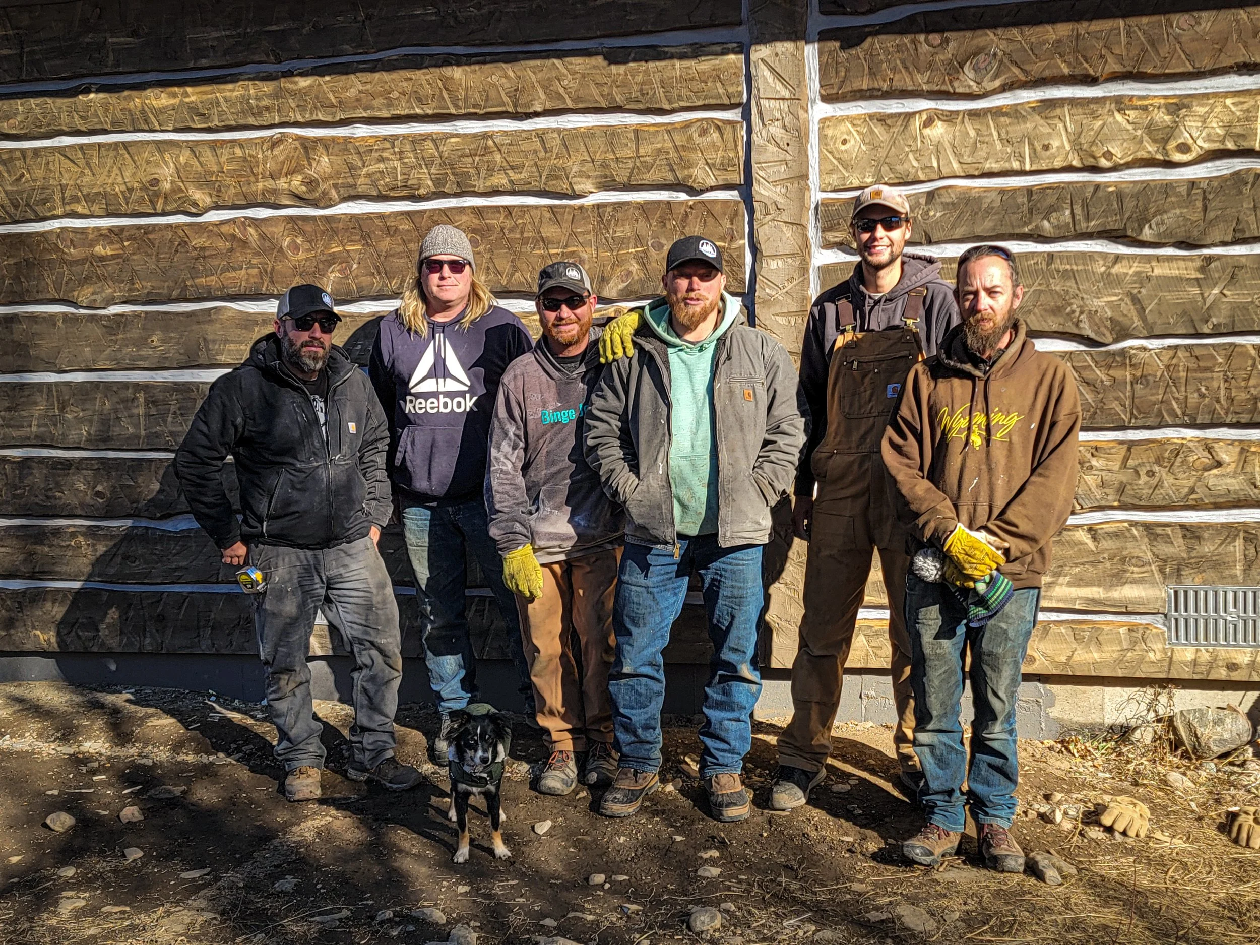 Six men and a dog standing in front of a wooden log cabin wall, dressed in work and outdoor clothing, on a rocky ground.
