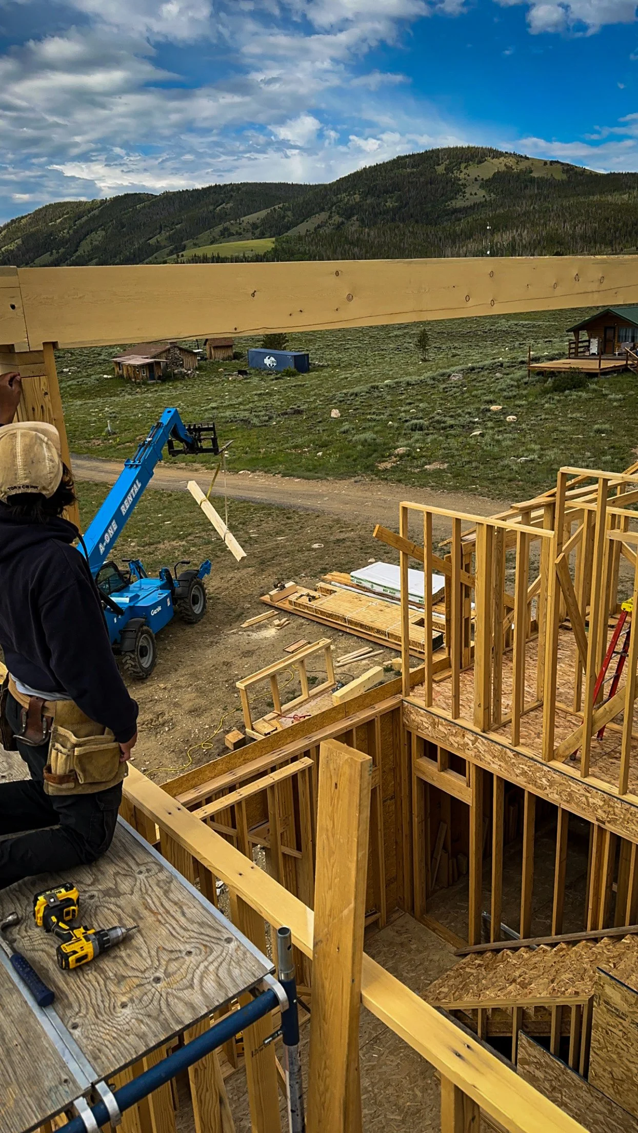 Construction site in progress with wooden framing for a building, a worker kneeling on a platform, tools such as a drill and a measuring tape visible, a blue crane lifting a piece of wood, and a scenic landscape with hills and a blue sky in the background.