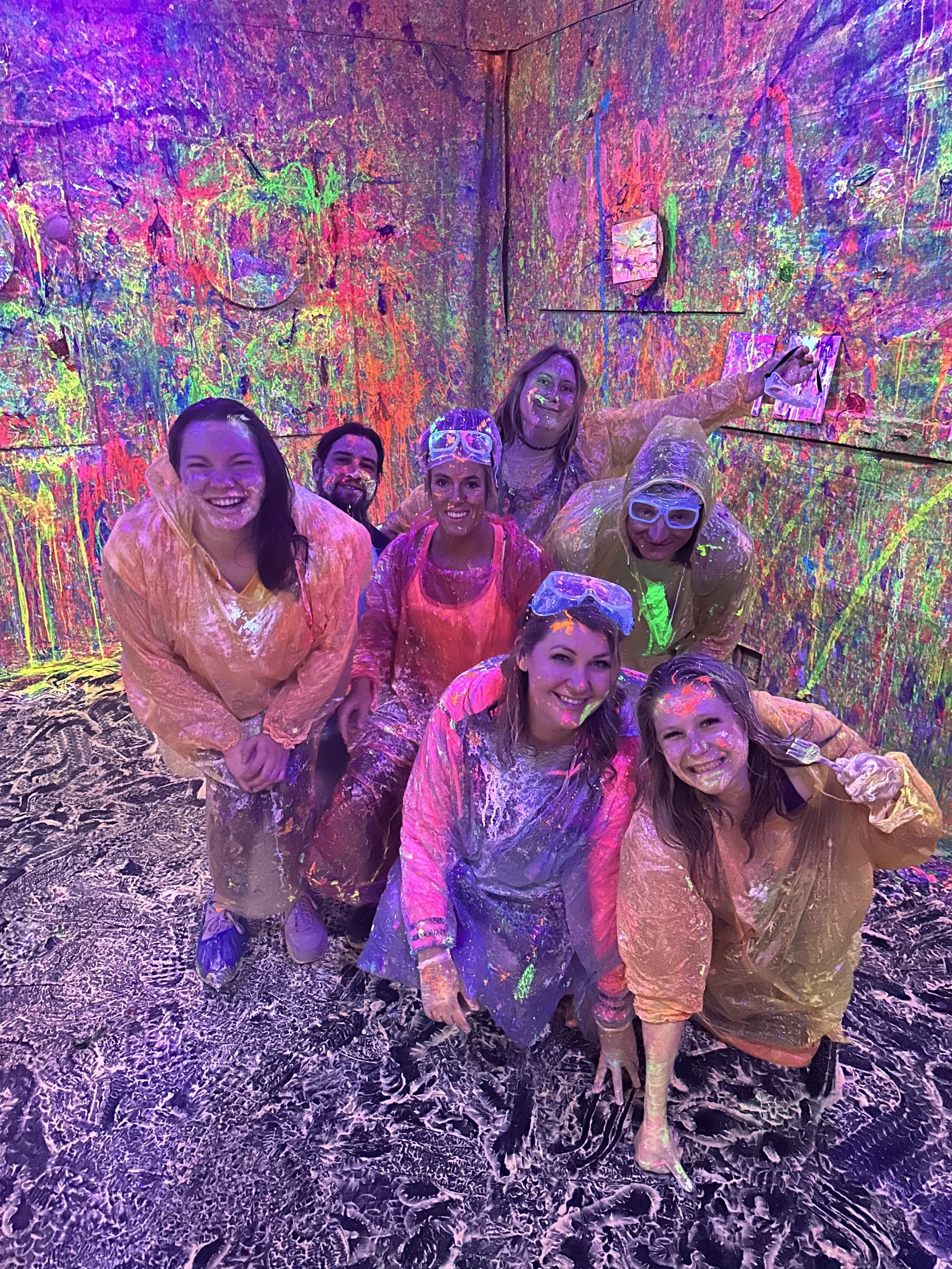 Group of happy people covered in colorful paint standing inside a room with walls and floor painted with bright paint splatters and designs.