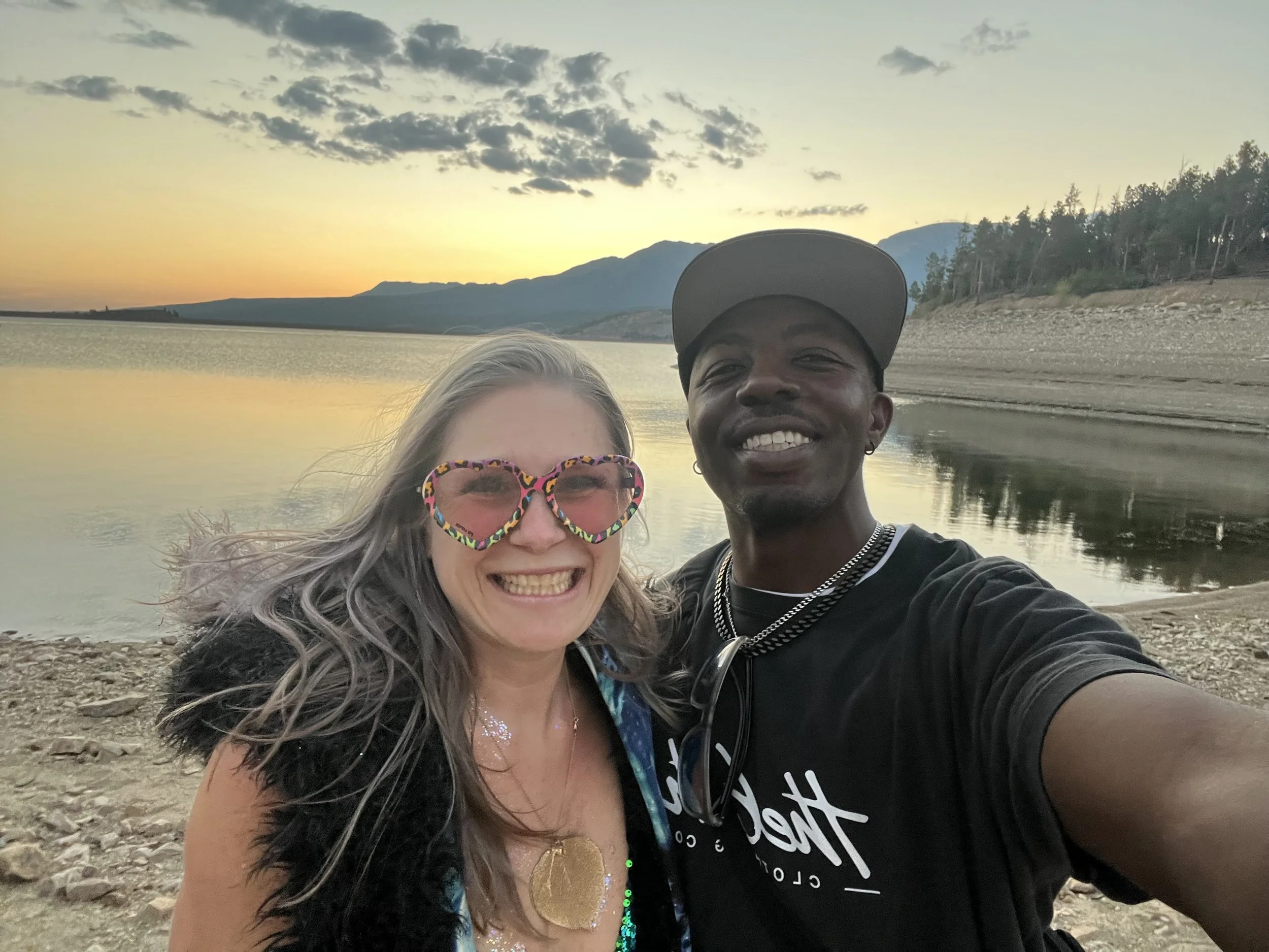 Photo of a smiling woman with sunglasses and a man taking a selfie by a body of water during a sunset, with mountains and trees in the background - Dillon, Colorado