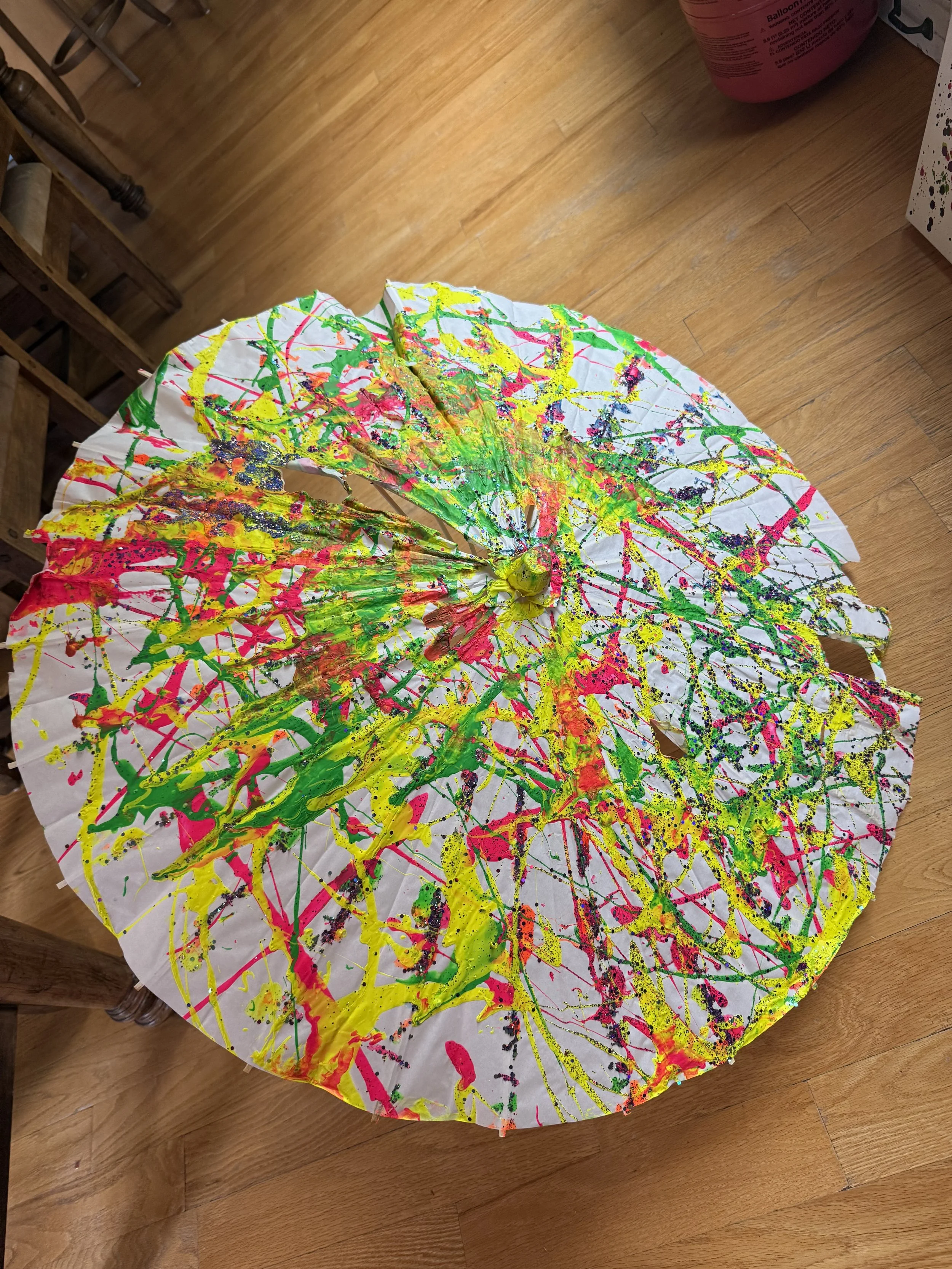 Colorful paper parasol with abstract splashes of yellow, pink, and green paint with glitter on a wooden floor.