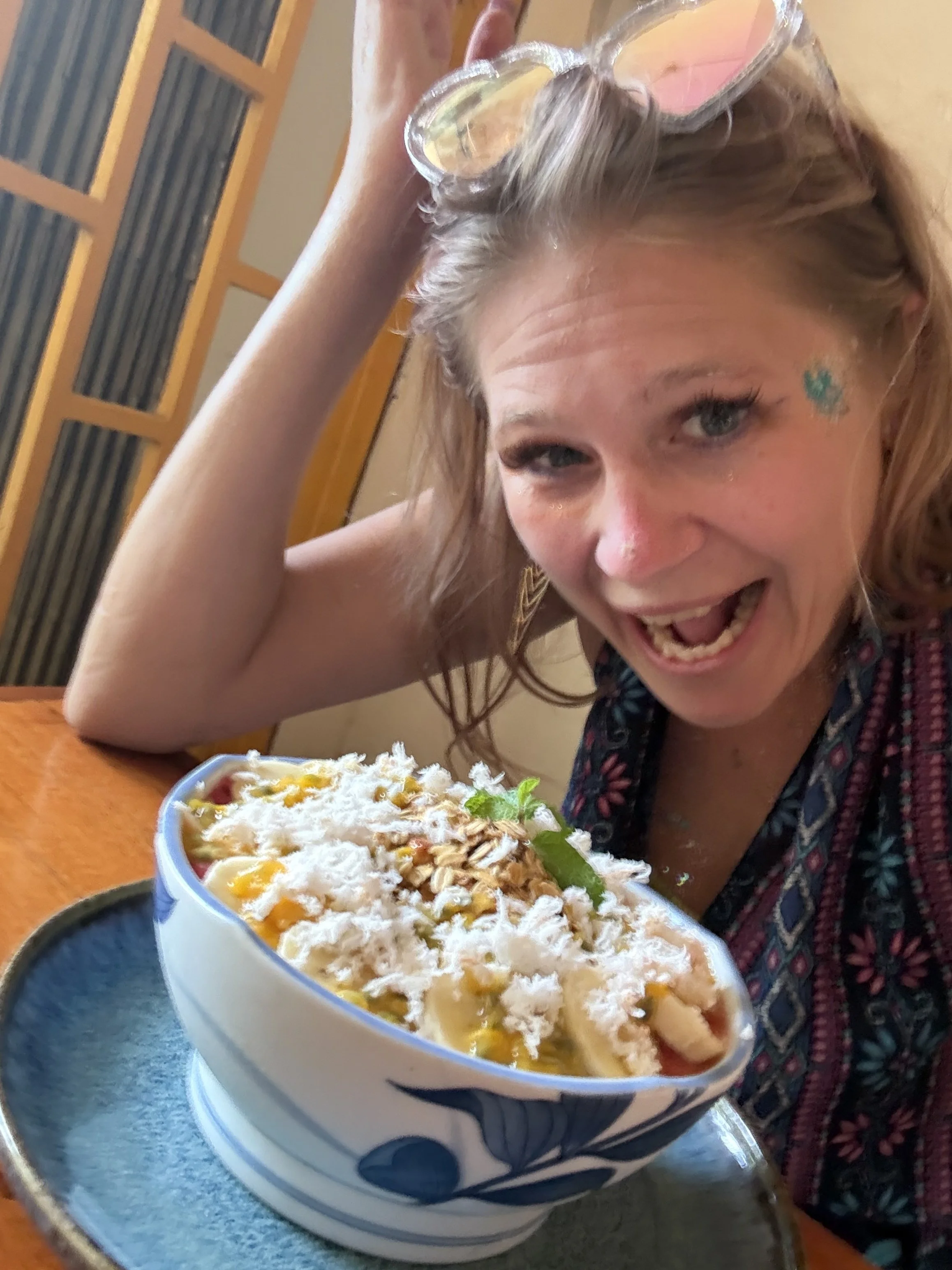 A young woman with blonde hair is smiling and appears excited about a large açai bowl with colorful tropical fruit and with shredded coconut, placed on a table.