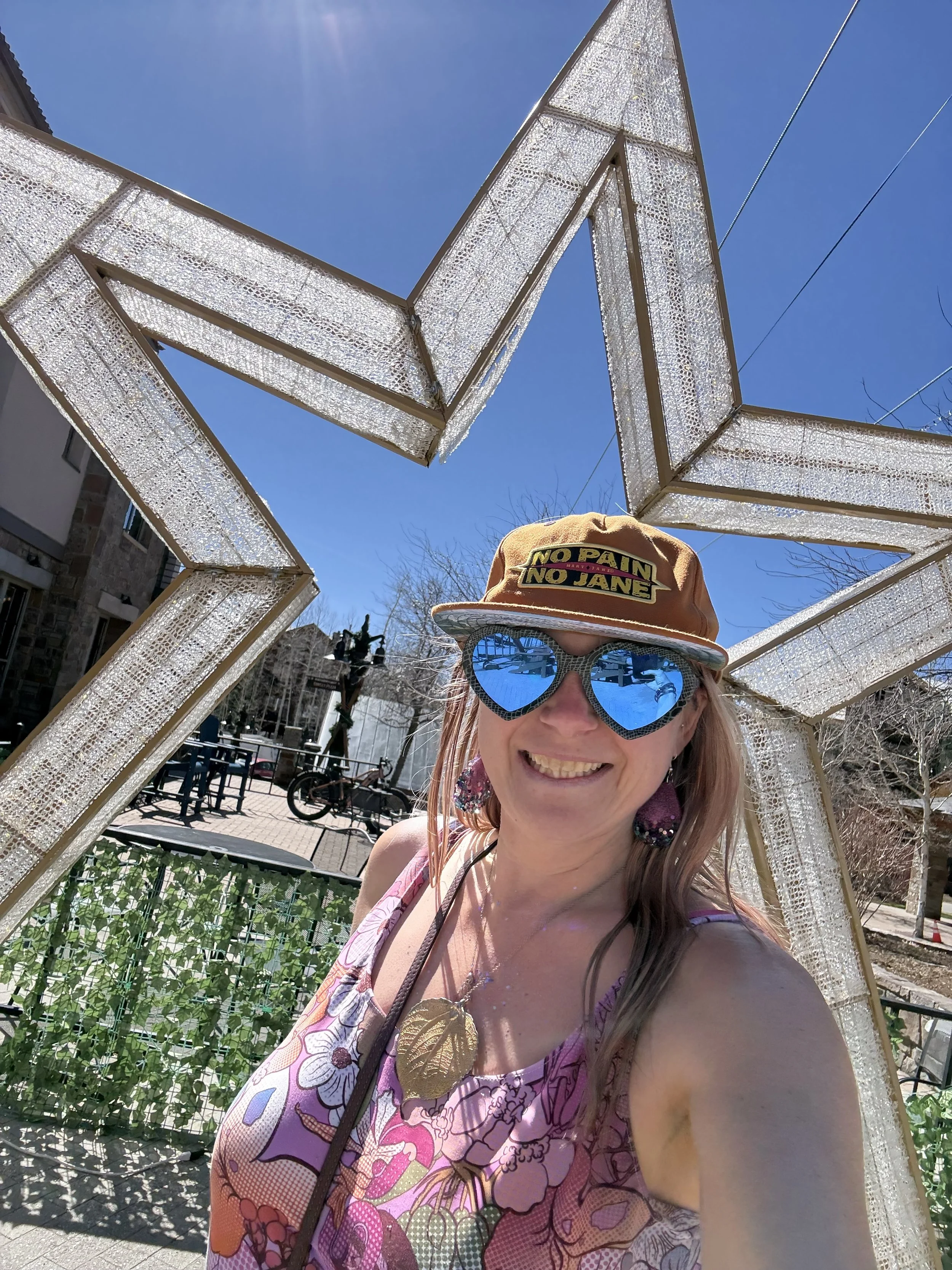 A woman smiling outdoors at a daytime event, wearing large blue heart-shaped sunglasses, a tan hat with a "No Pain No Jane" patch, and a pink floral dress, standing under a decorative structure with a bright blue sky background.