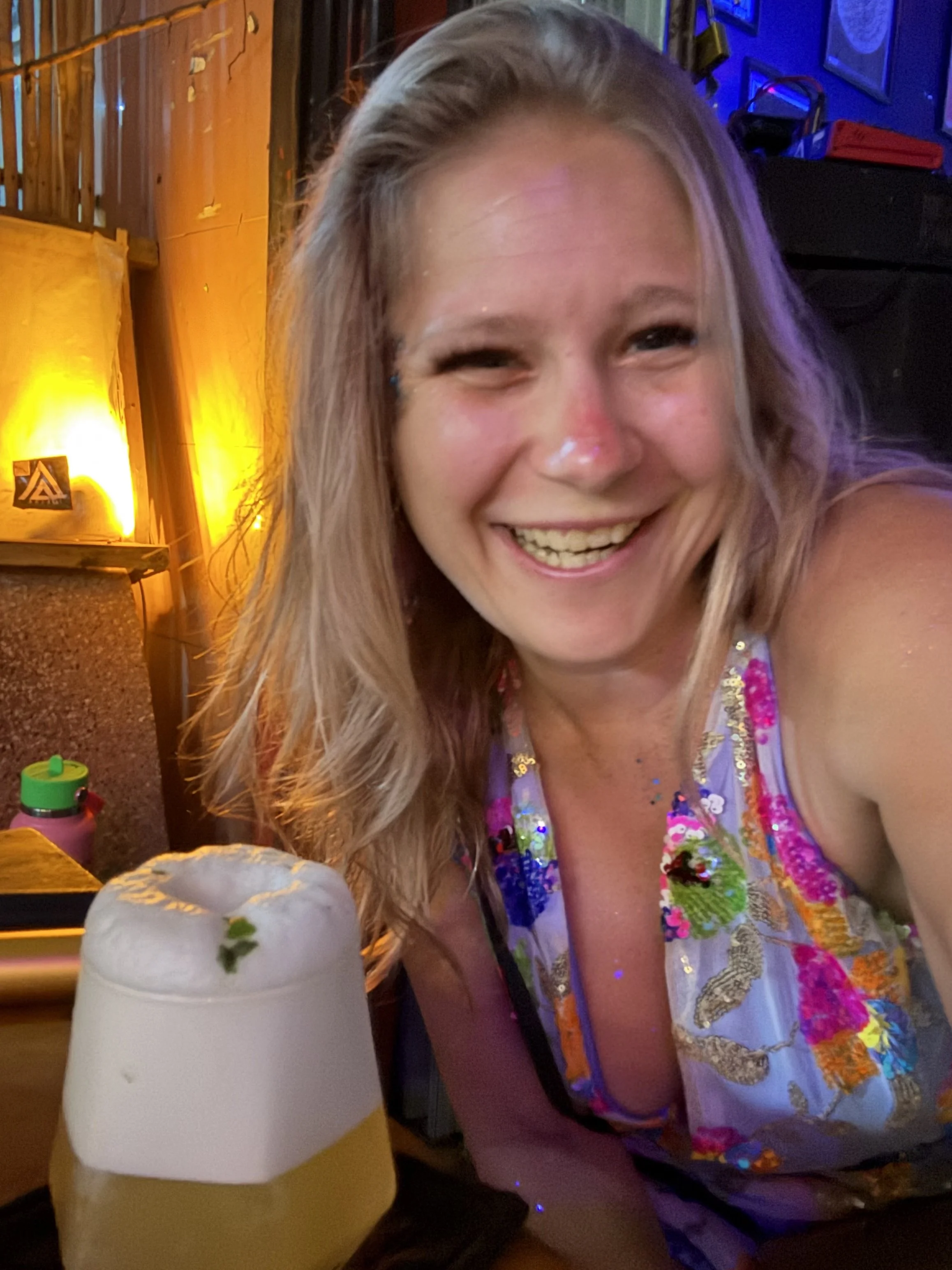 A woman with long blonde hair smiling at a bar, wearing a colorful dress. In front of her is a frothy whiskey sour with a lime wedge on the rim of the glass.