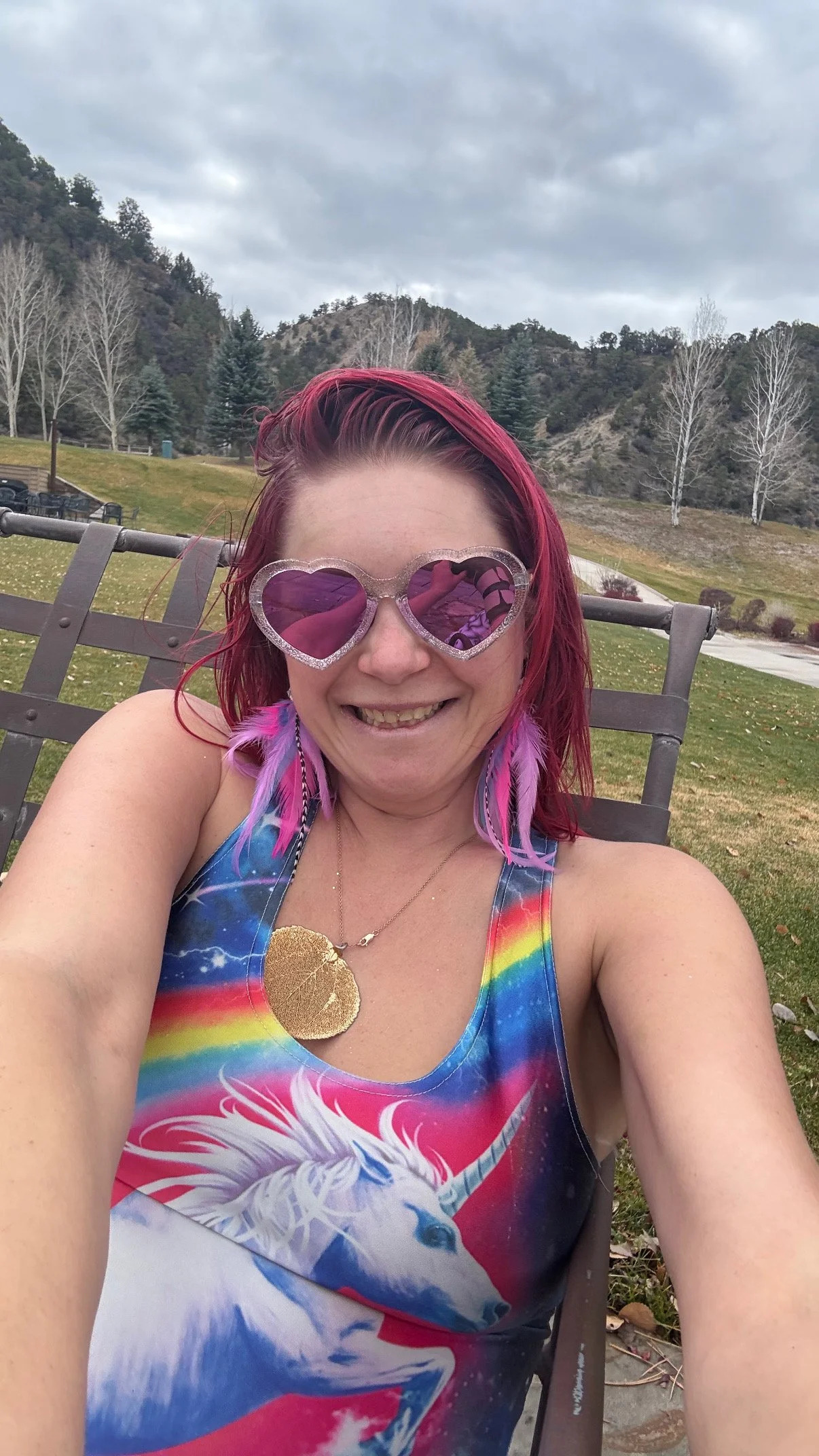 A woman with pink hair wearing heart-shaped pink sunglasses and a rainbow-colored unicorn themed dress, smiling while sitting outside on a bench with a background of hills, trees, and cloudy sky.