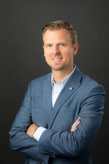 A man with short, light brown hair and blue eyes wearing a blue blazer over a white dress shirt, standing against a dark background.