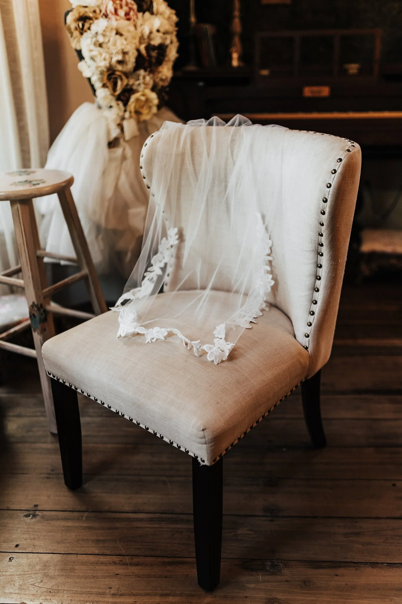 A beige upholstered chair with dark wooden legs, decorated with a bridal veil featuring floral embroidery, in a room with wooden floors and vintage furniture.