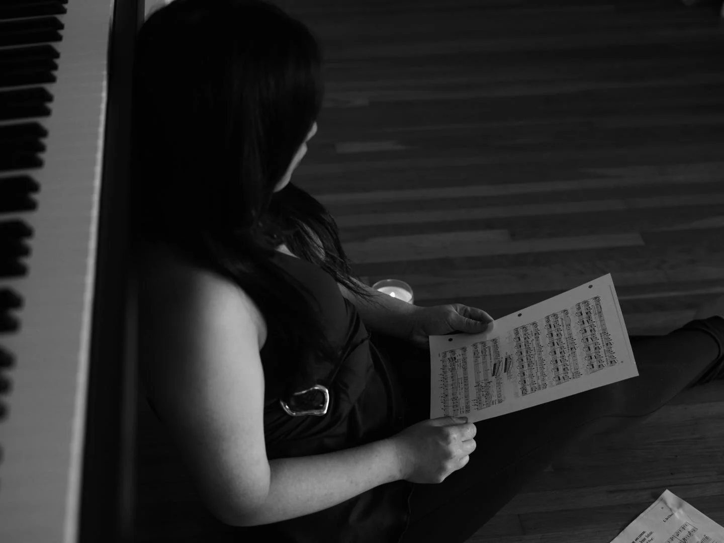A woman sitting on a wooden floor, reading sheet music, with a piano keyboard partly visible on the left and a candle glowing nearby.