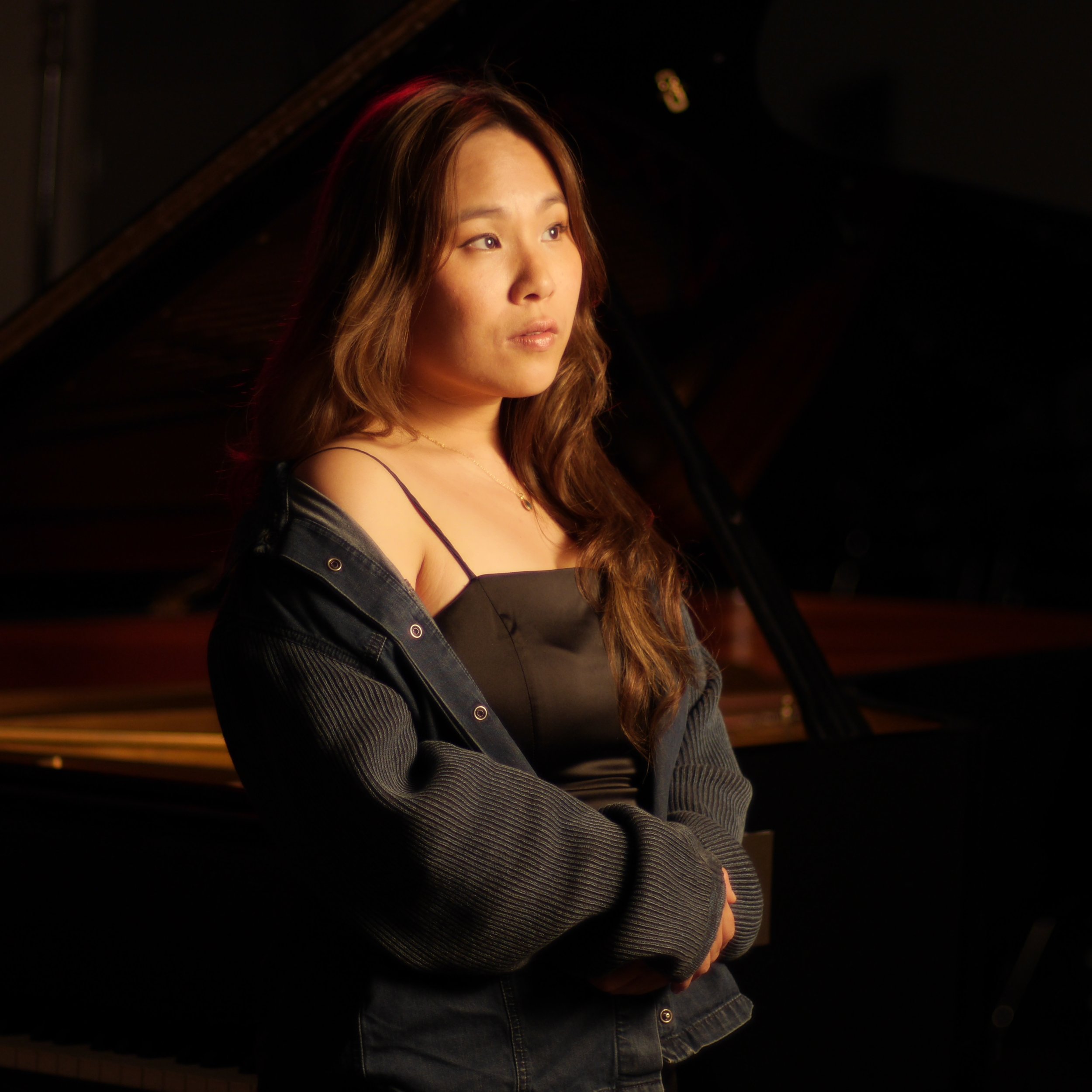 Young woman with long brown hair, wearing a black satin dress with thin straps and a dark jacket, standing in front of a grand piano in a dimly lit setting.