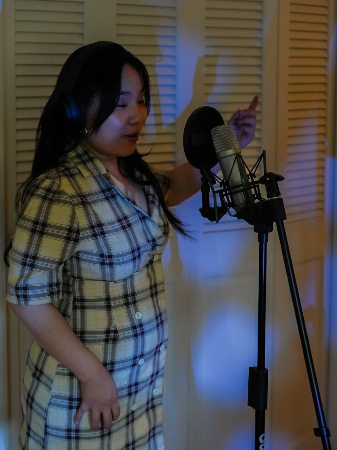 A woman with long dark hair wearing a plaid dress singing into a studio microphone in a recording booth.