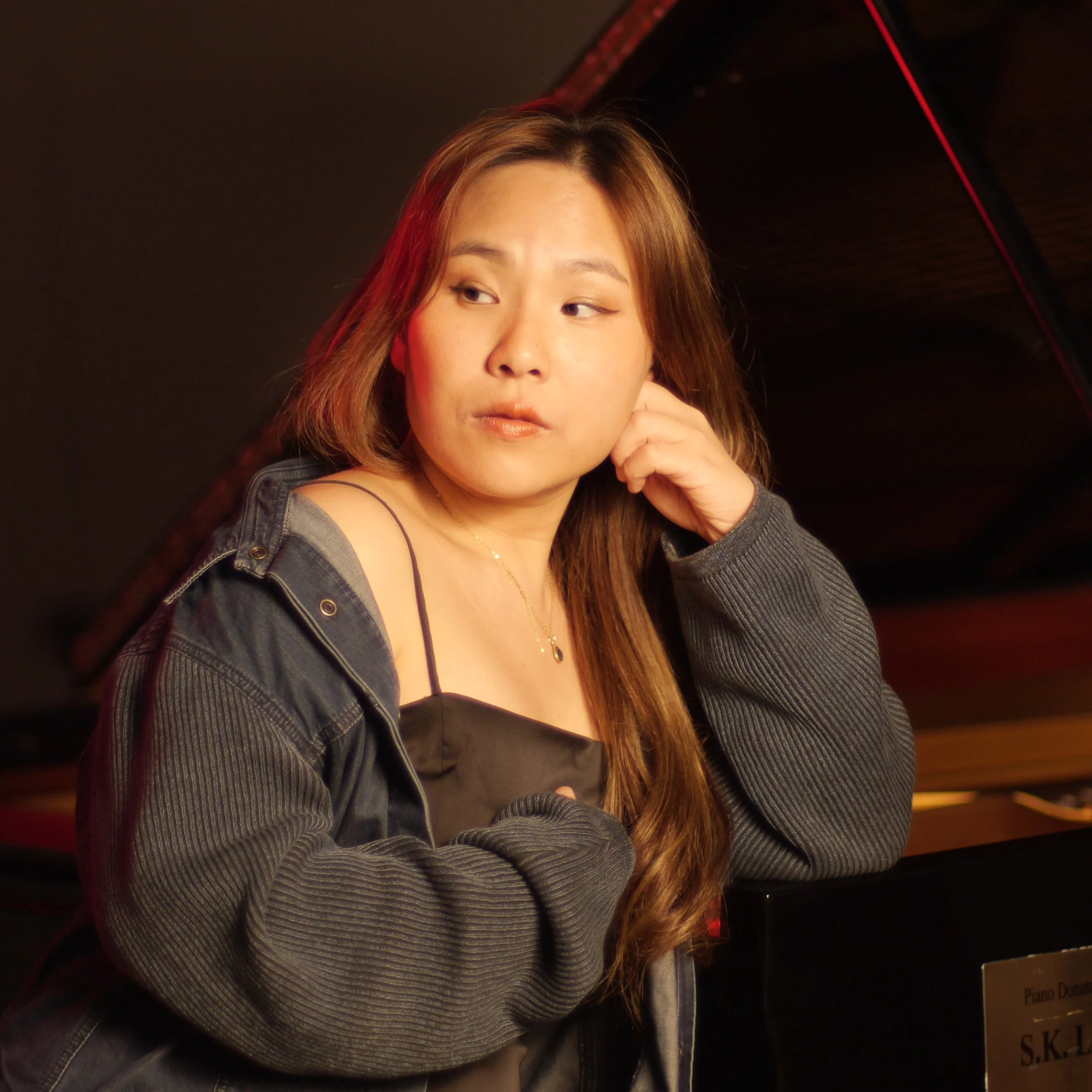 A young woman with light brown hair and a black top leaning against a piano with a dark background.