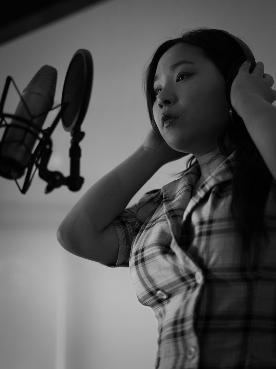 A woman in plaid shirt wearing headphones, recording vocals in a sound studio with a microphone.