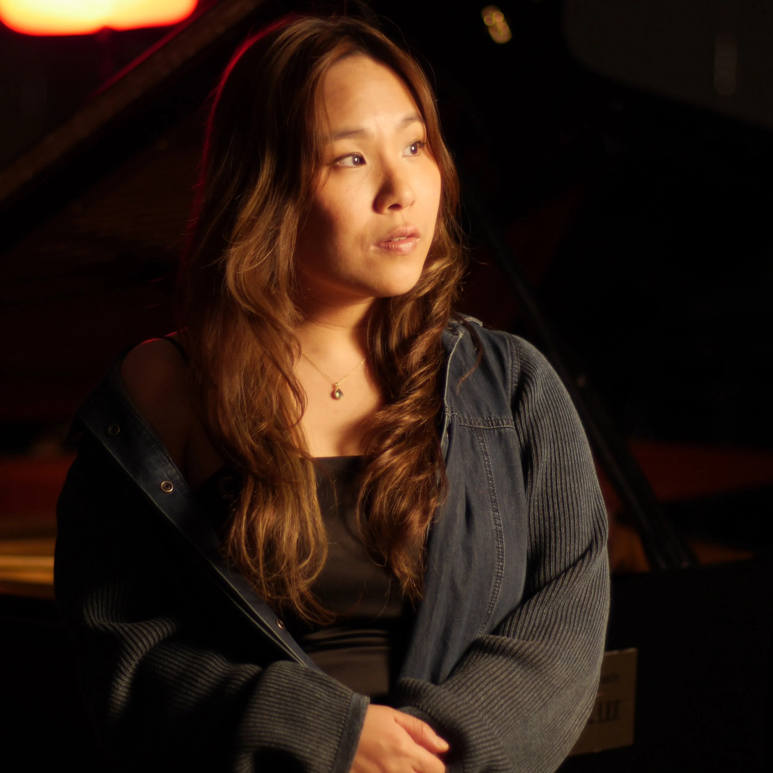 A young woman with long wavy hair wearing a black top and gray jacket, sitting near a piano, looking thoughtfully to the side in low lighting.