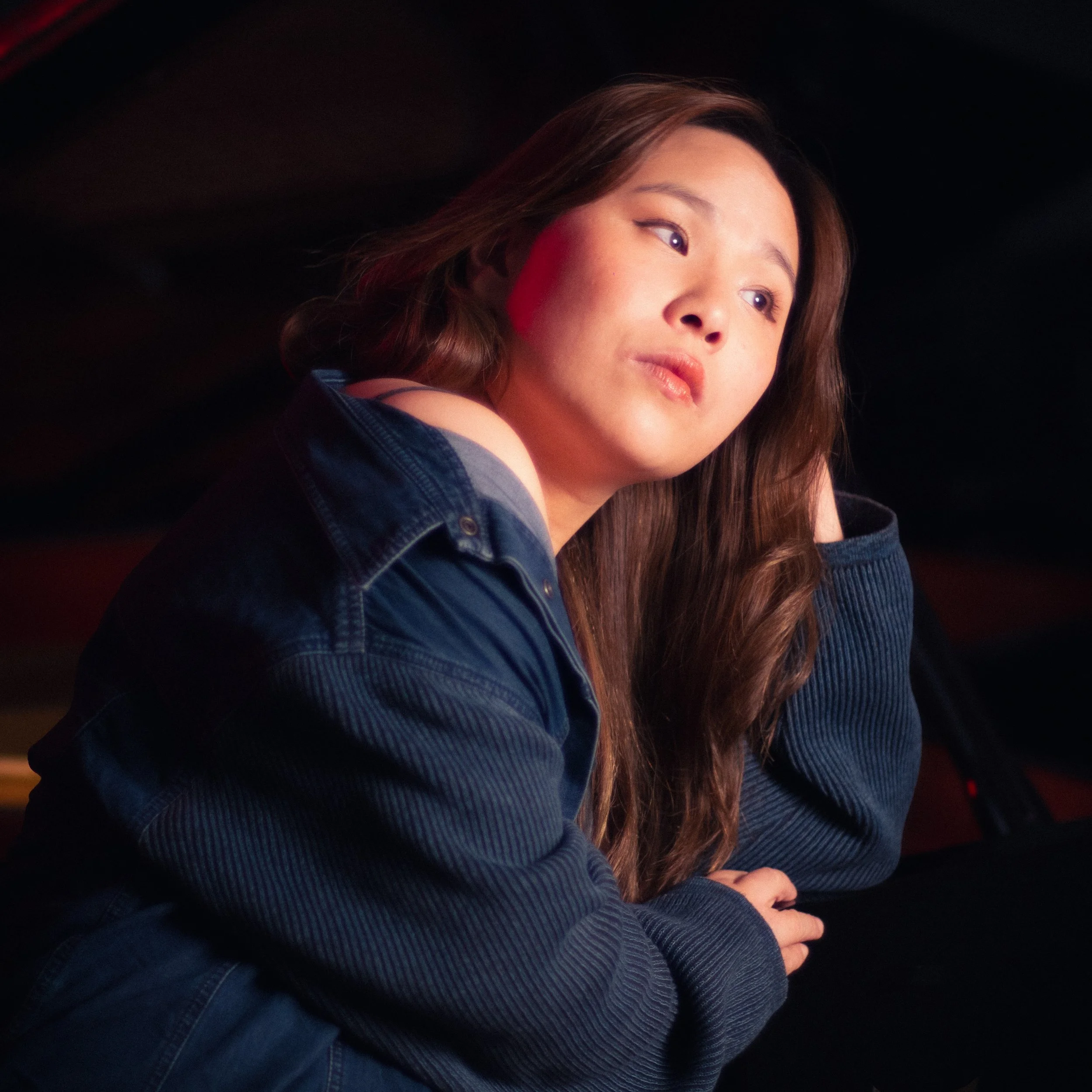 A young woman with long brown hair wearing a denim jacket resting her head on her hand in a dimly lit setting.