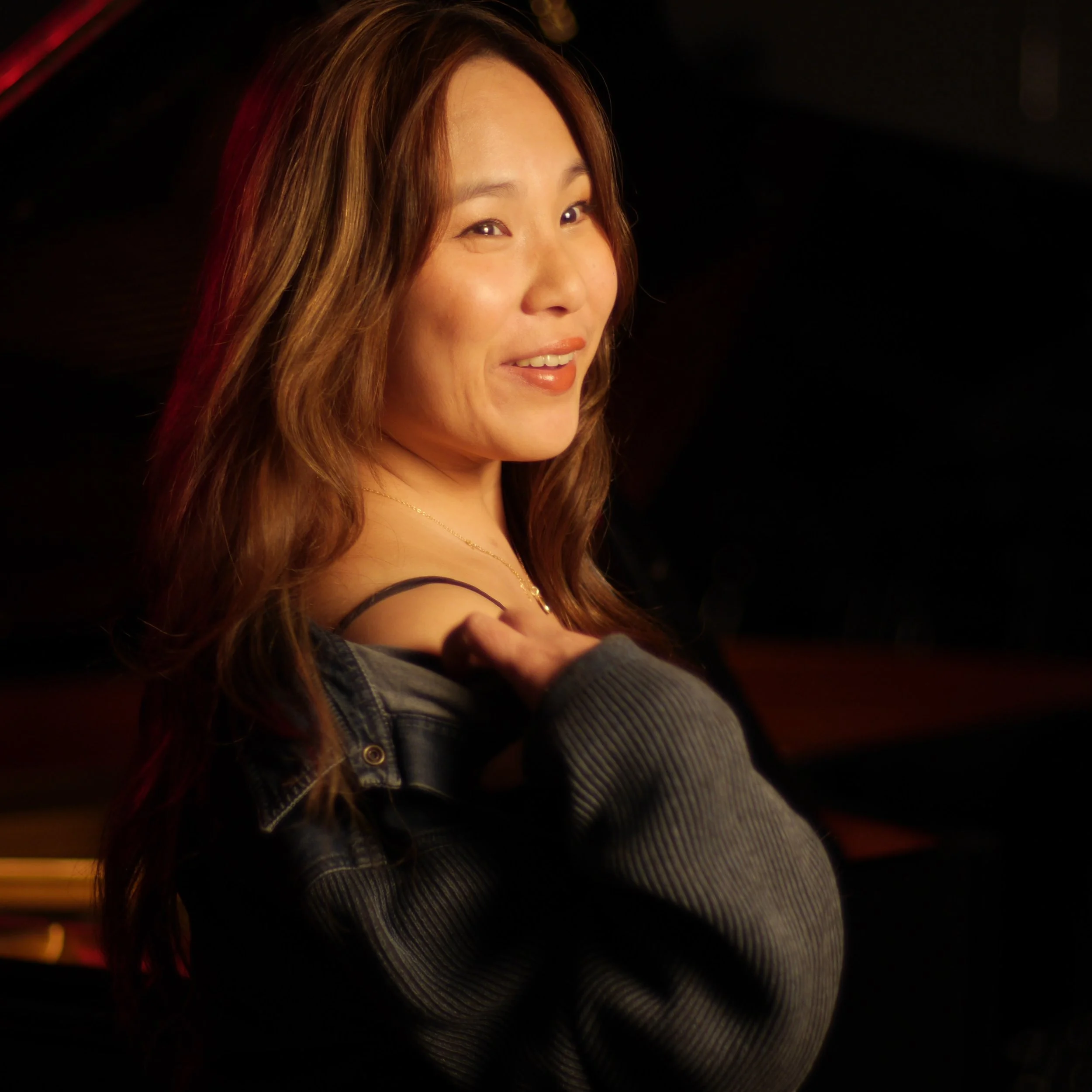 A woman with long wavy hair smiling at the camera, wearing a dark off-the-shoulder top and a gray sweater.