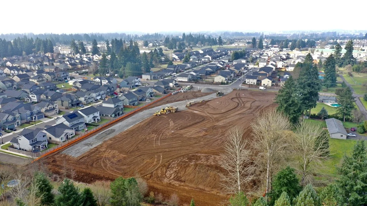A construction site with dirt and earth-moving equipment surrounded by a residential neighborhood with houses, trees, and a swimming pool.
