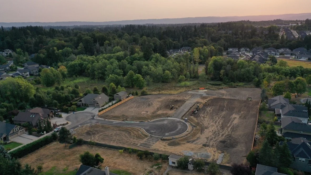 A residential neighborhood with several houses surrounding a large under-construction plot of land, cleared and leveled, with construction equipment present and a circular driveway area.