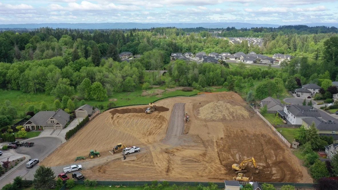 Construction site with multiple heavy machinery, including bulldozers and trucks, leveling and grading land in a residential neighborhood surrounded by lush green trees.