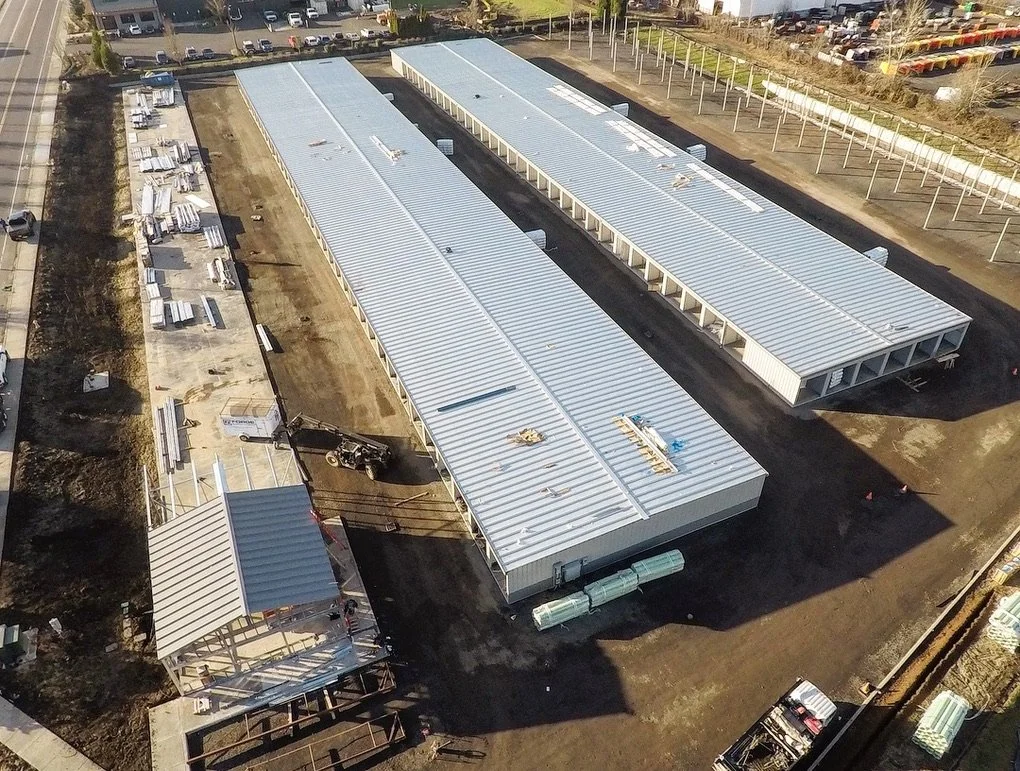 Aerial view of a construction site with two large rectangular warehouse buildings with metal roofs, a small building with a porch, and construction materials and equipment around the site.