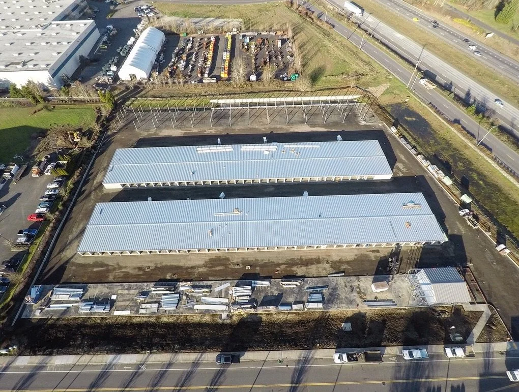Aerial view of a construction site with two long buildings with metal roofs, dirt surrounding them, and construction materials in the front. A parking lot with vehicles, a fenced area, and a highway with traffic are visible in the background.